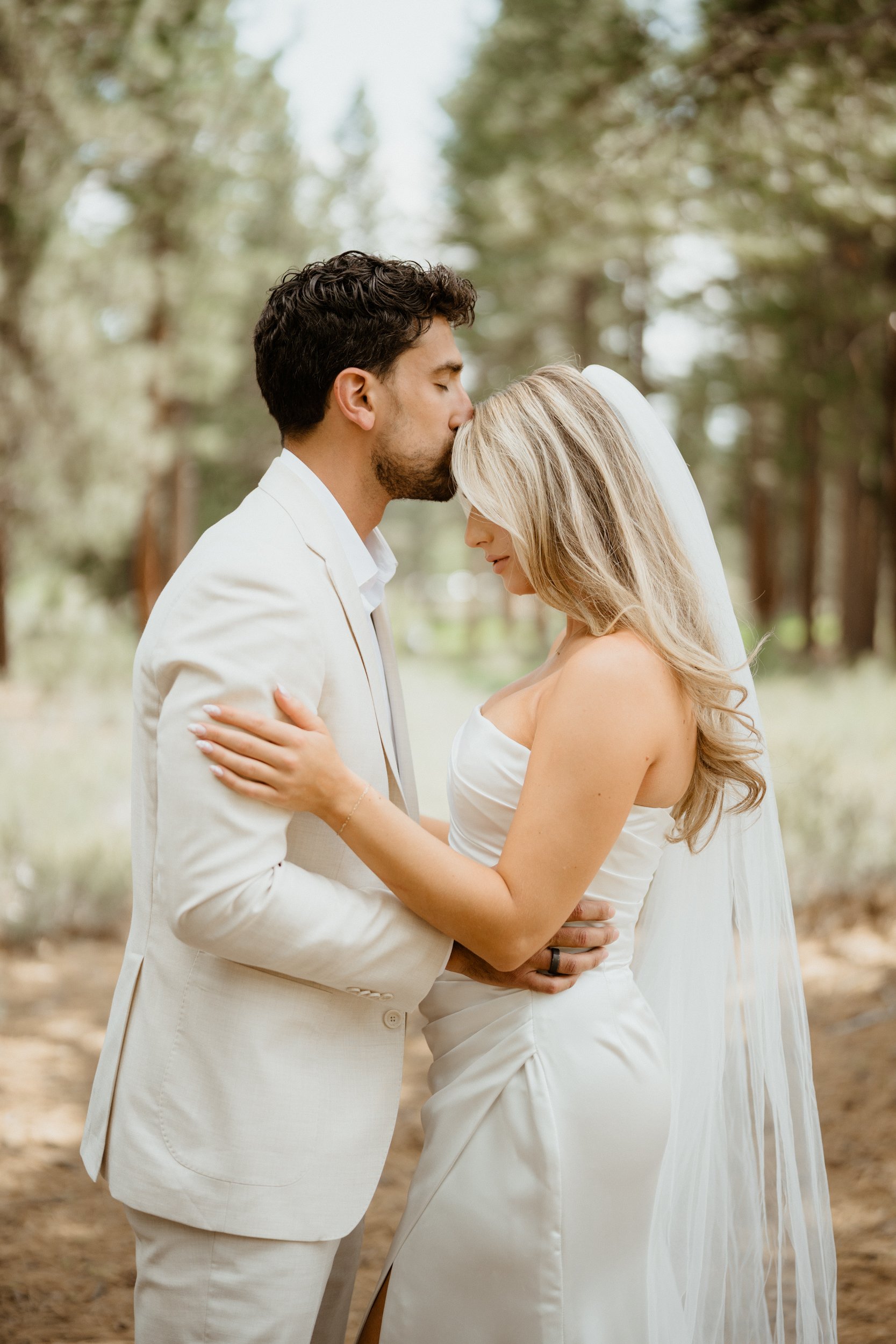 A couple in wedding attire embracing outdoors, with the man kissing the woman's forehead in a forest setting.