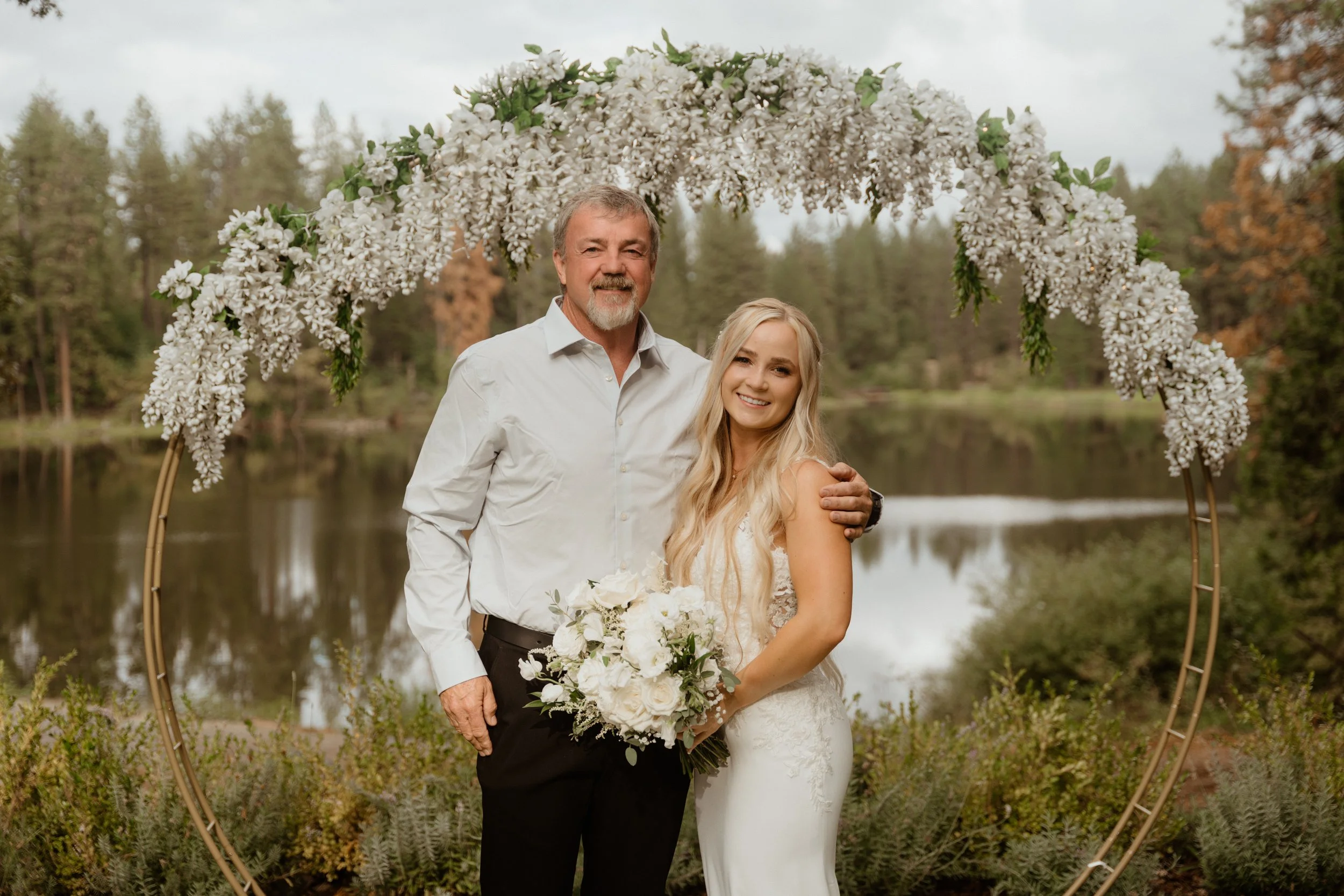 A man and woman in wedding attire standing under a circular floral arch outdoors near a lake, with trees in the background. The woman holds a bouquet of white flowers, and they are smiling.