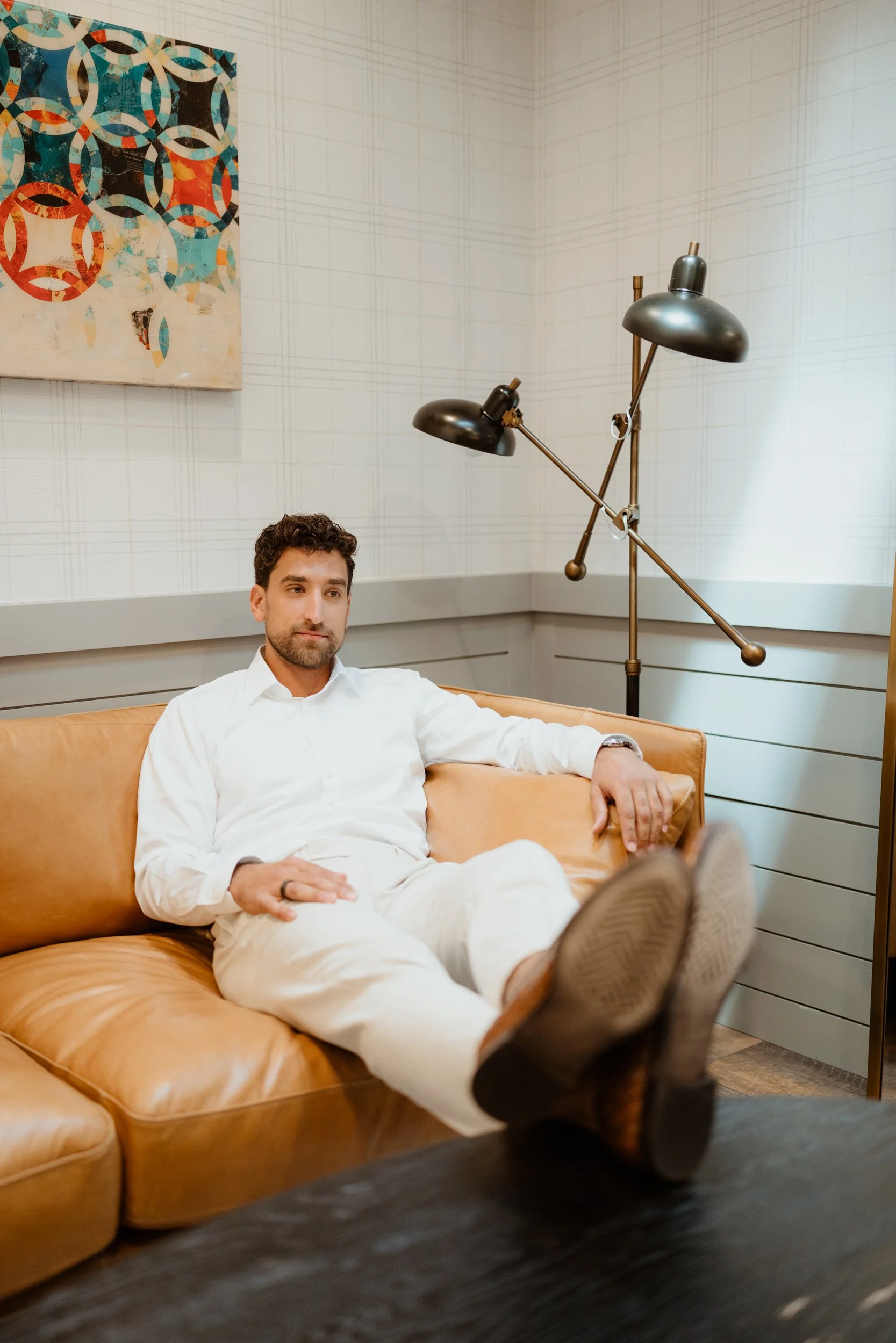Man sitting on a tan leather sofa with feet up, wearing a white shirt and pants, in a room with modern decor and a colorful abstract painting on the white wall.