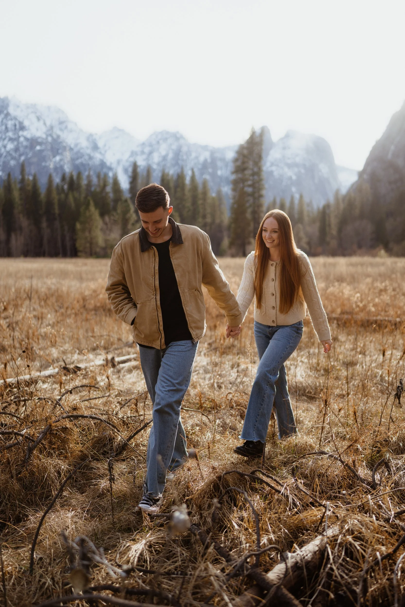 A young couple walking hand in hand through a meadow with tall grass and fallen branches, surrounded by a forest with mountain peaks in the background, under bright sky.