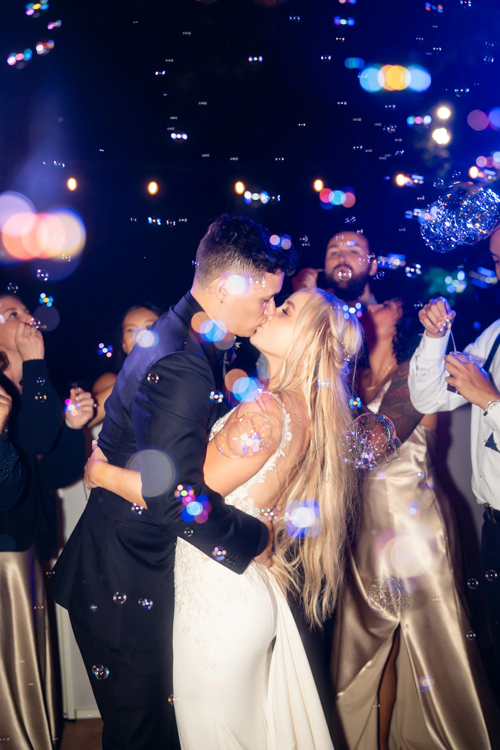 Couple kissing during a wedding celebration surrounded by friends, with bubbles floating in the air and colorful lights creating a festive atmosphere.