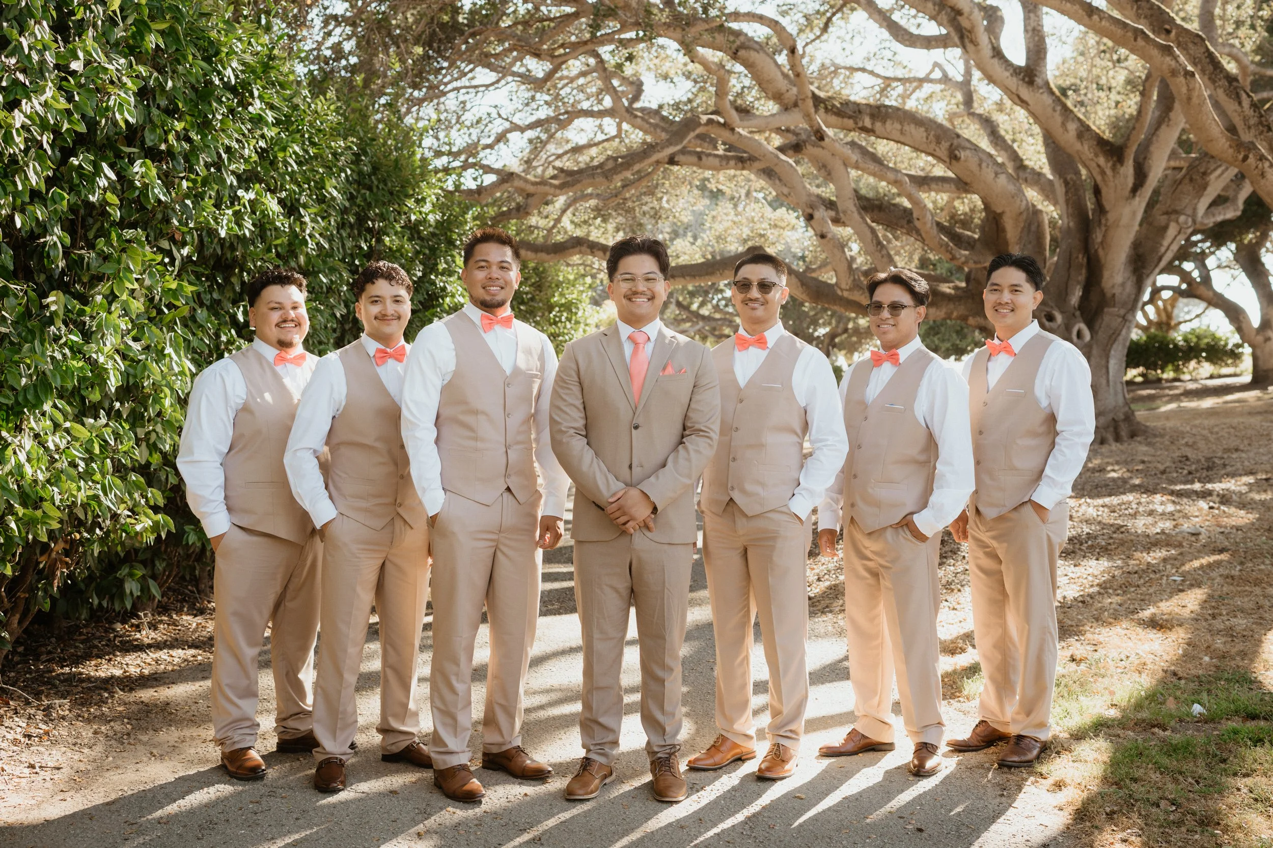A group of eight men wearing matching beige suits, white shirts, and pink bow ties, standing outdoors on a sunny day with trees in the background, smiling at the camera.