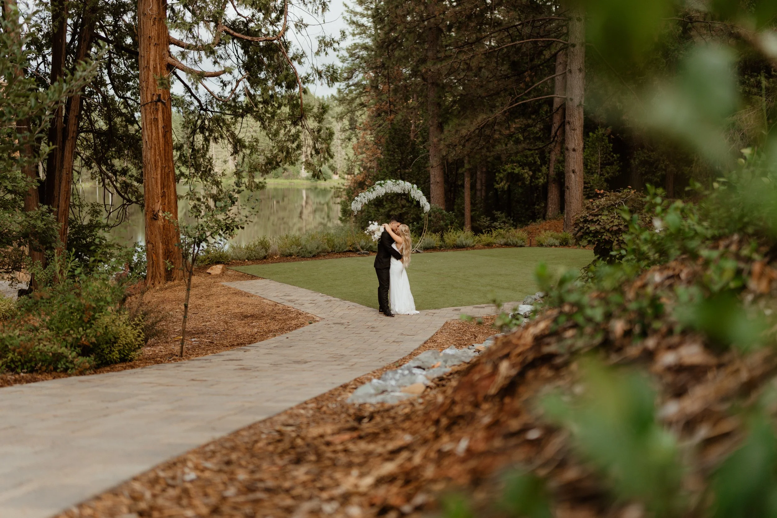 A bride and groom embrace and kiss on a pathway in a park near a lake surrounded by trees, with a decorative floral arch in the background.