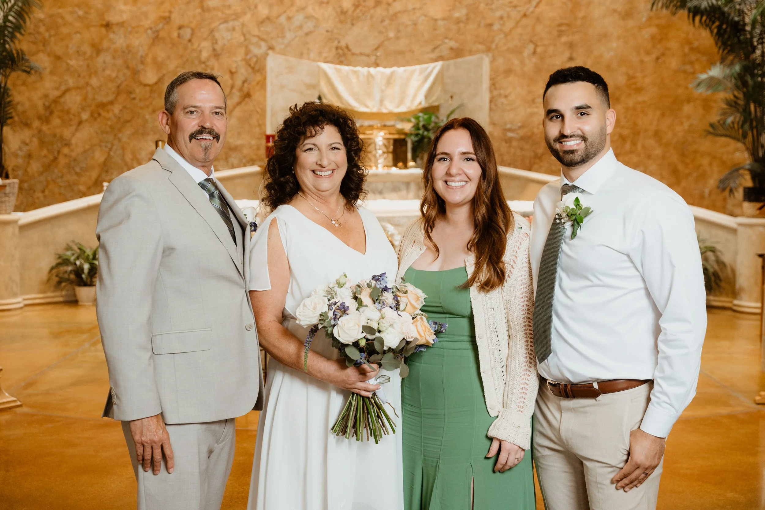 A group of four people, two men and two women, standing together indoors in front of a decorative backdrop, smiling for a photo. The woman second from the left is holding a bouquet of flowers.