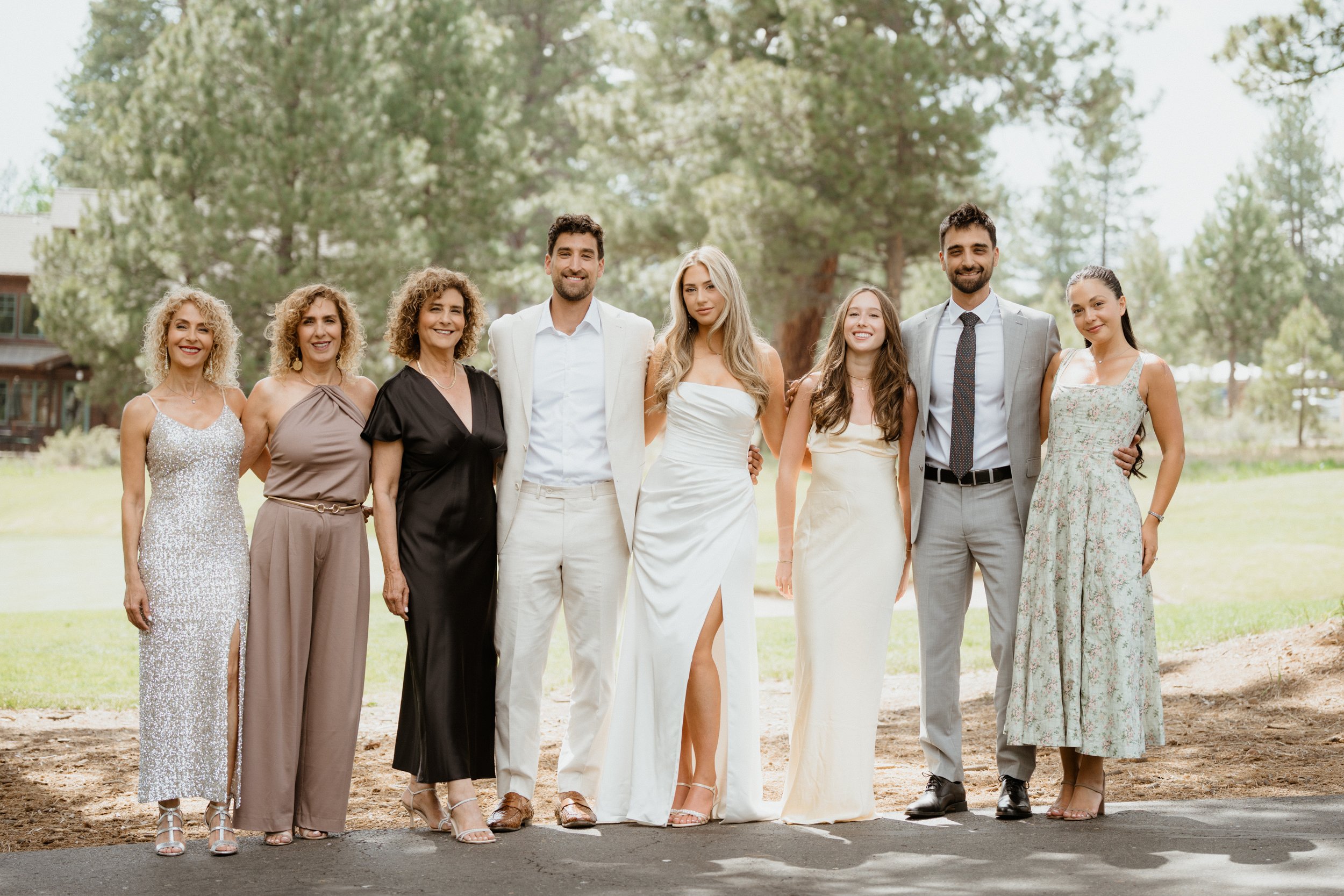 Group of people standing outdoors, dressed in formal attire, smiling, with trees and a house in the background.