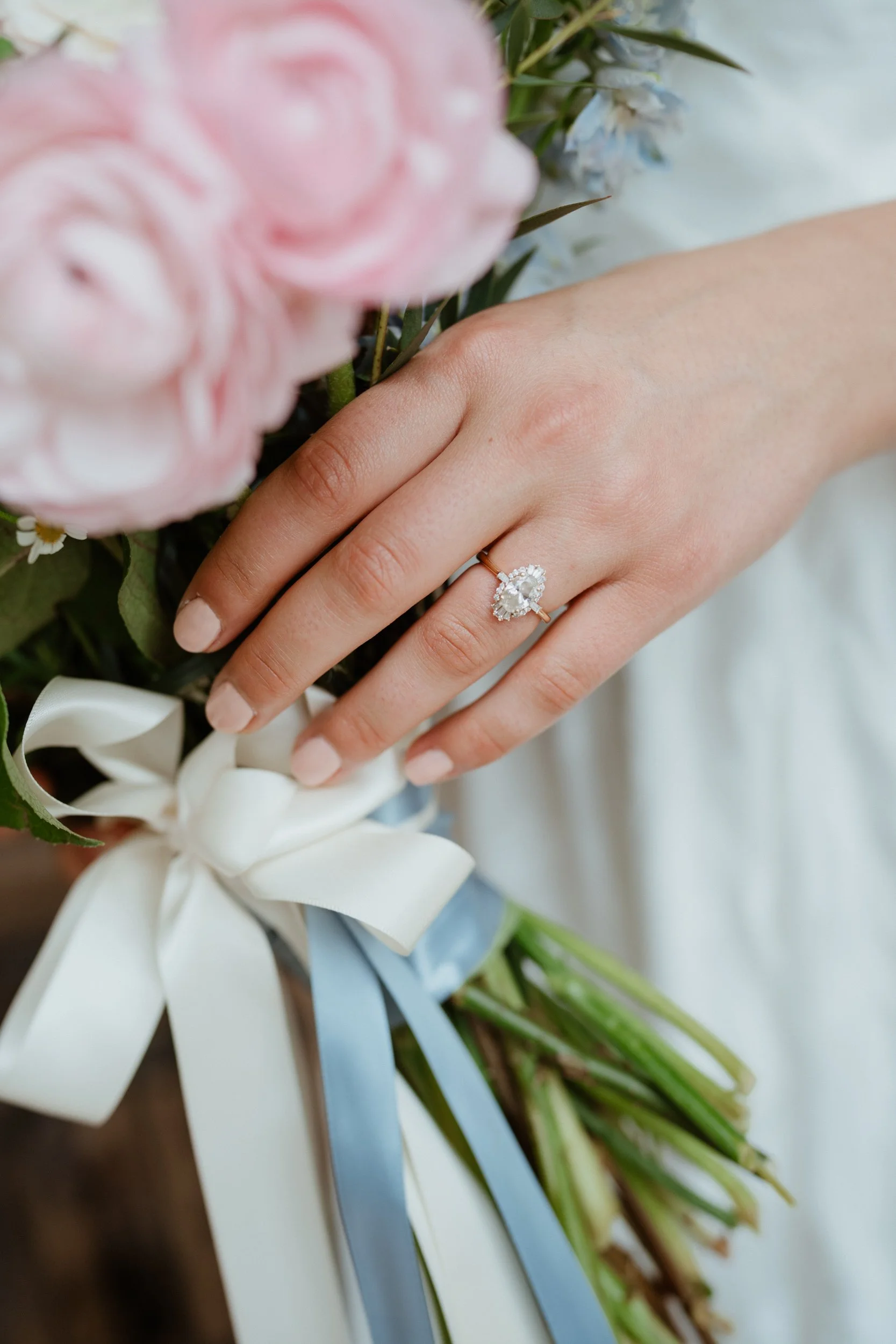 Close-up of a woman's hand showing an engagement ring, holding a bouquet of pink and white flowers tied with white and blue ribbons.