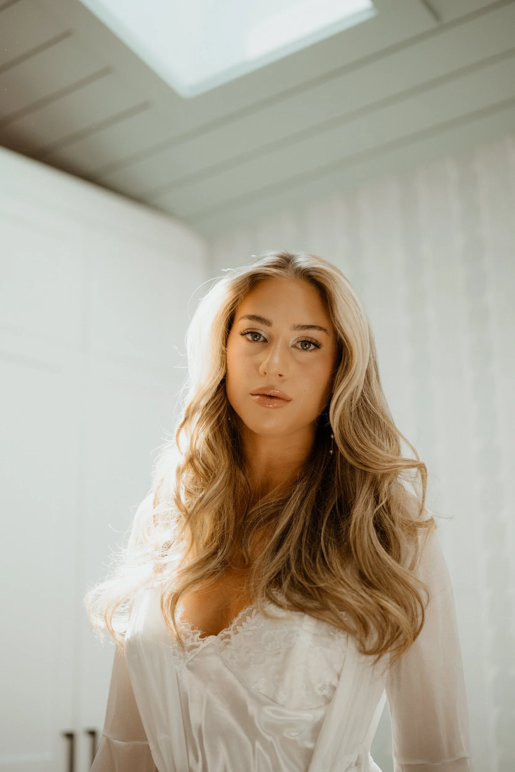A woman with long, wavy blonde hair and makeup, wearing a white satin or silk top with lace details, standing in a bright, minimally decorated room with a white wall and ceiling.