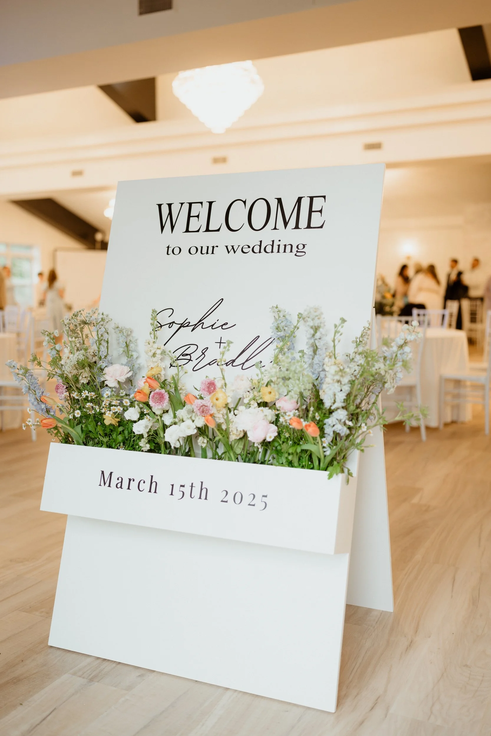 Wedding welcome sign with floral decorations, displaying names 'Sophie & Bradley' and date March 15th, 2025, inside a decorated venue.