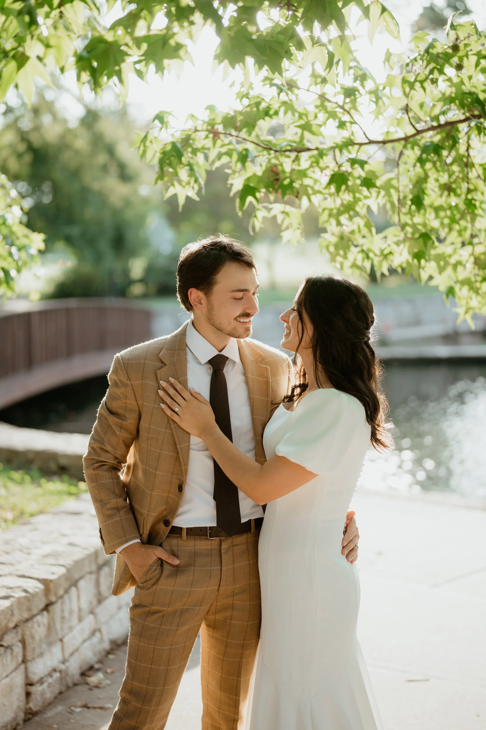 A couple dressed in wedding attire standing outdoors under green leafy trees near a small water body, smiling at each other.