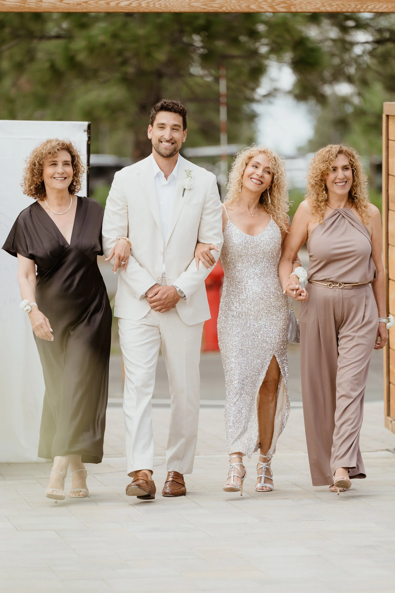 Group of four women and one man dressed in formal attire walking outdoors, smiling, with trees in the background.