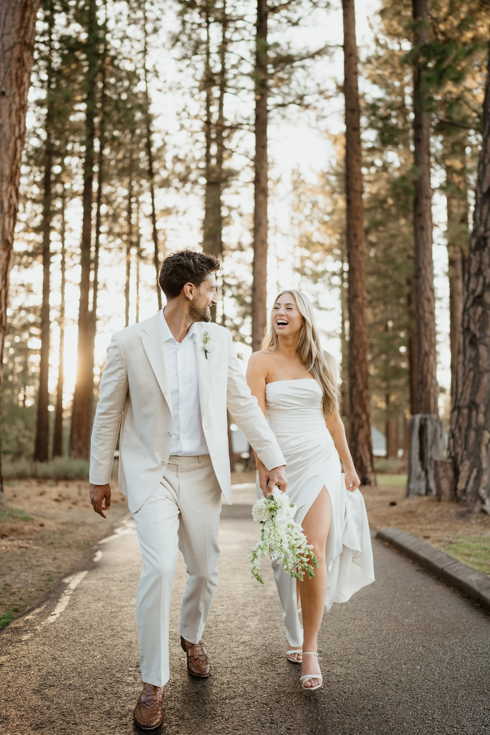 A bride and groom walking hand in hand through a wooded area, smiling and laughing, during their wedding day sunset.