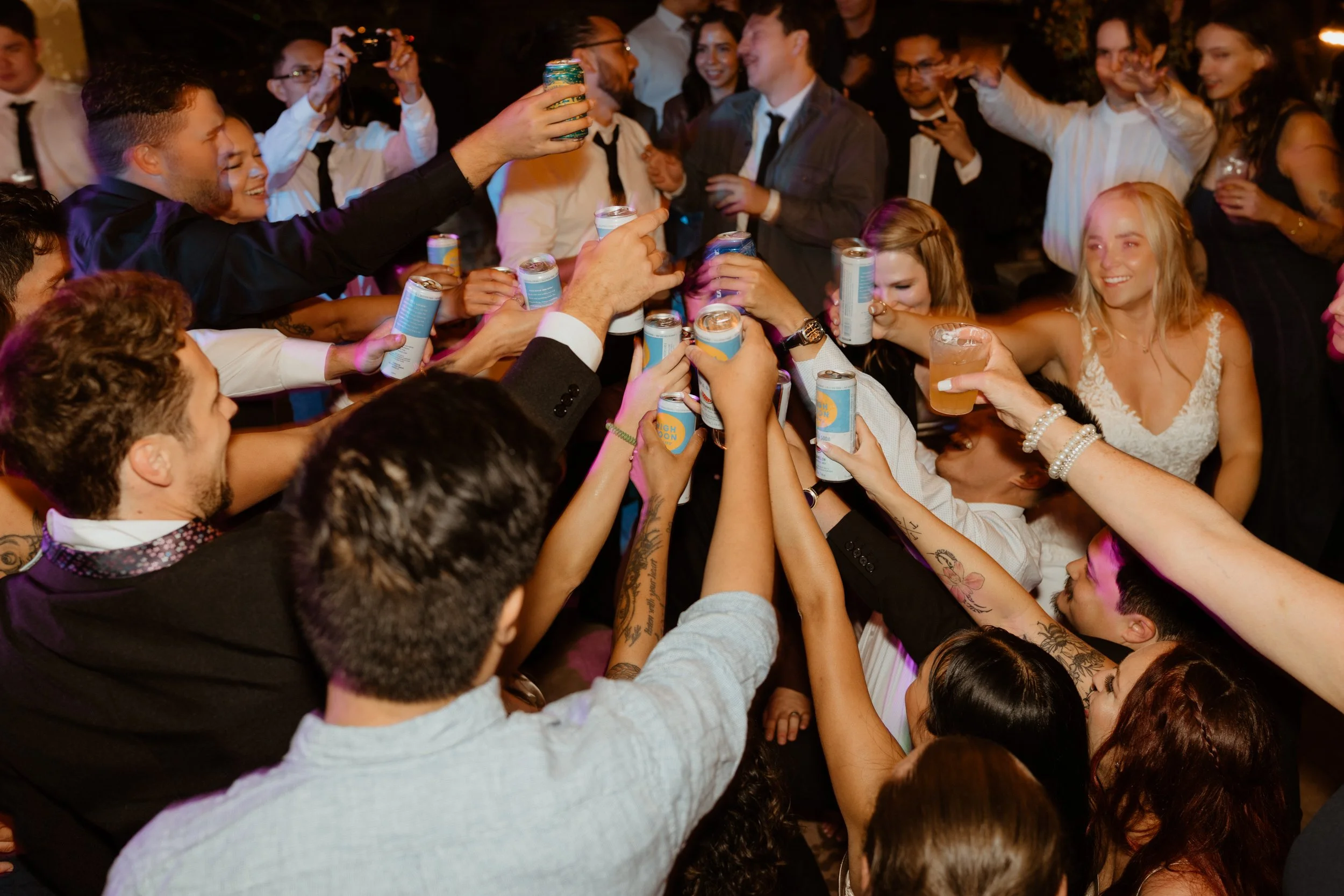 Group of people raising cans and glasses in a toast at a party, with smiling faces and various drinks.