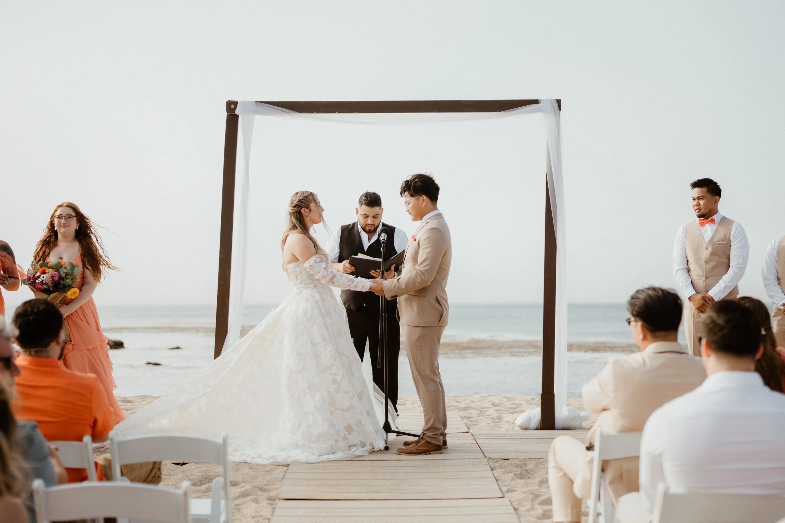 A beach wedding ceremony with a bride and groom holding hands under a wooden arch, with an officiant reading from a book. Guests are seated on white chairs, and a bridesmaid holds a bouquet. The background shows the ocean and sky.
