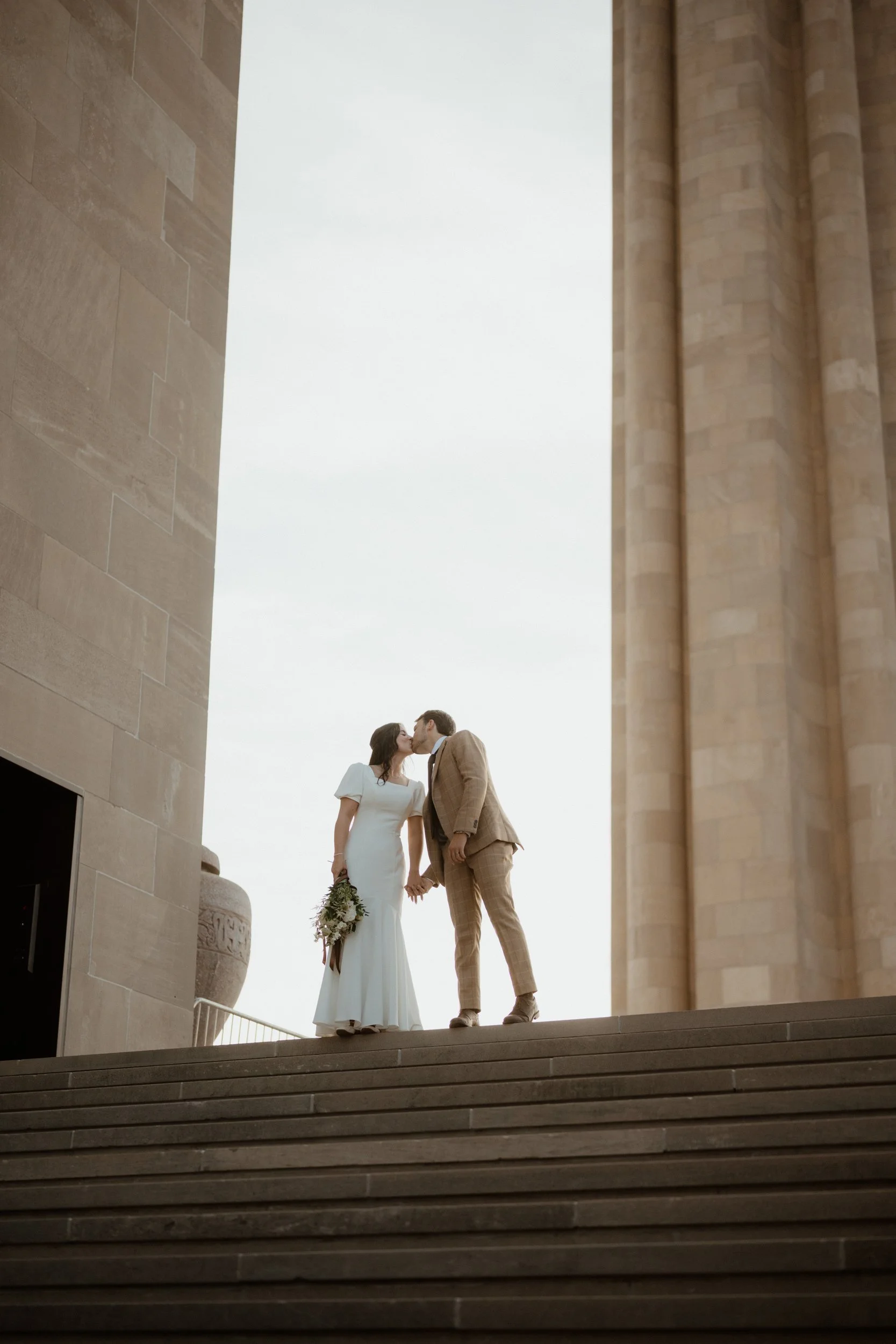 A bride and groom kiss on the steps outside a beige stone building, holding hands, with the bride holding a bouquet of flowers.