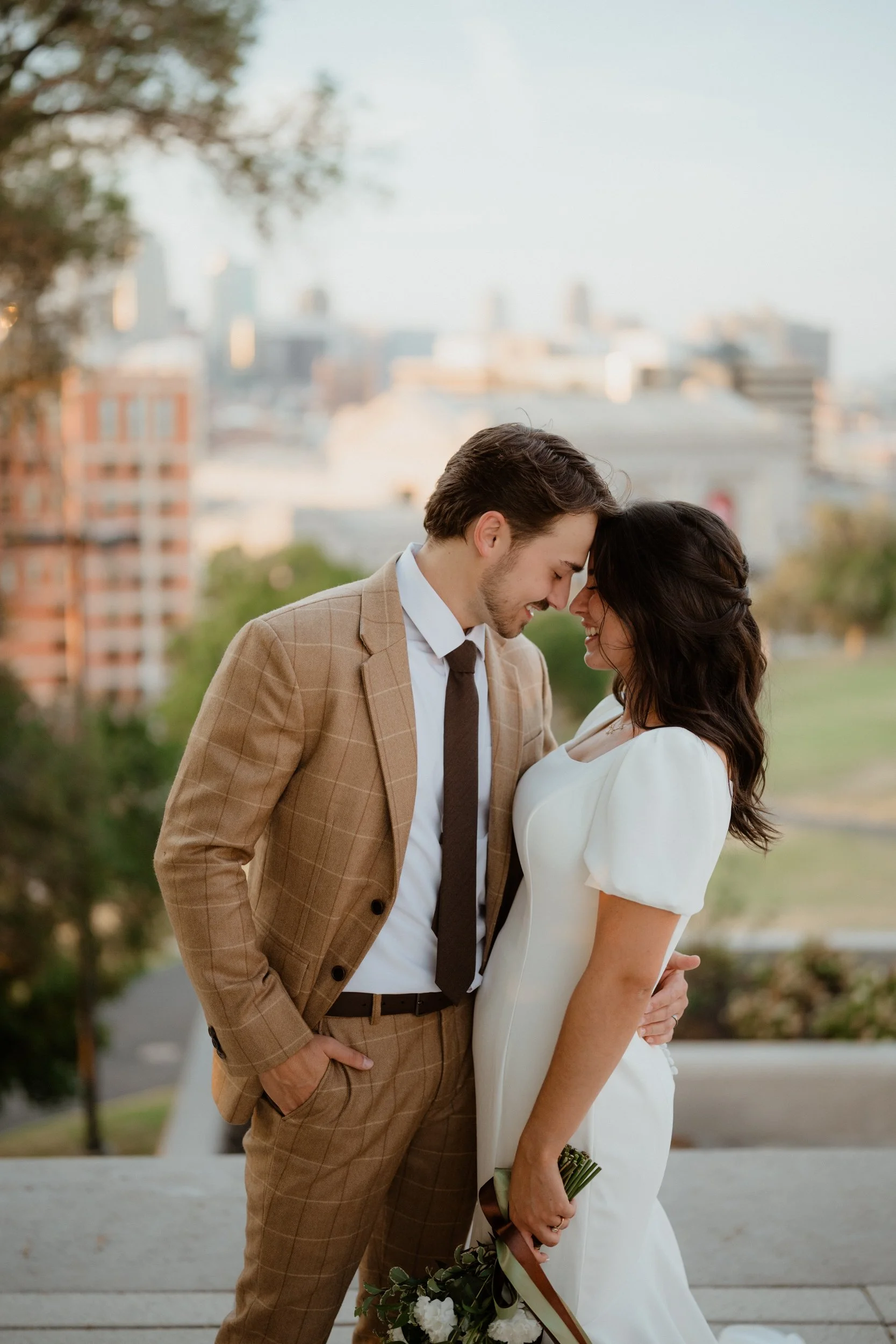 A couple sharing a tender moment on their wedding day, with foreheads touching and eyes closed, holding a bouquet of white flowers outdoors in a city park at sunset.