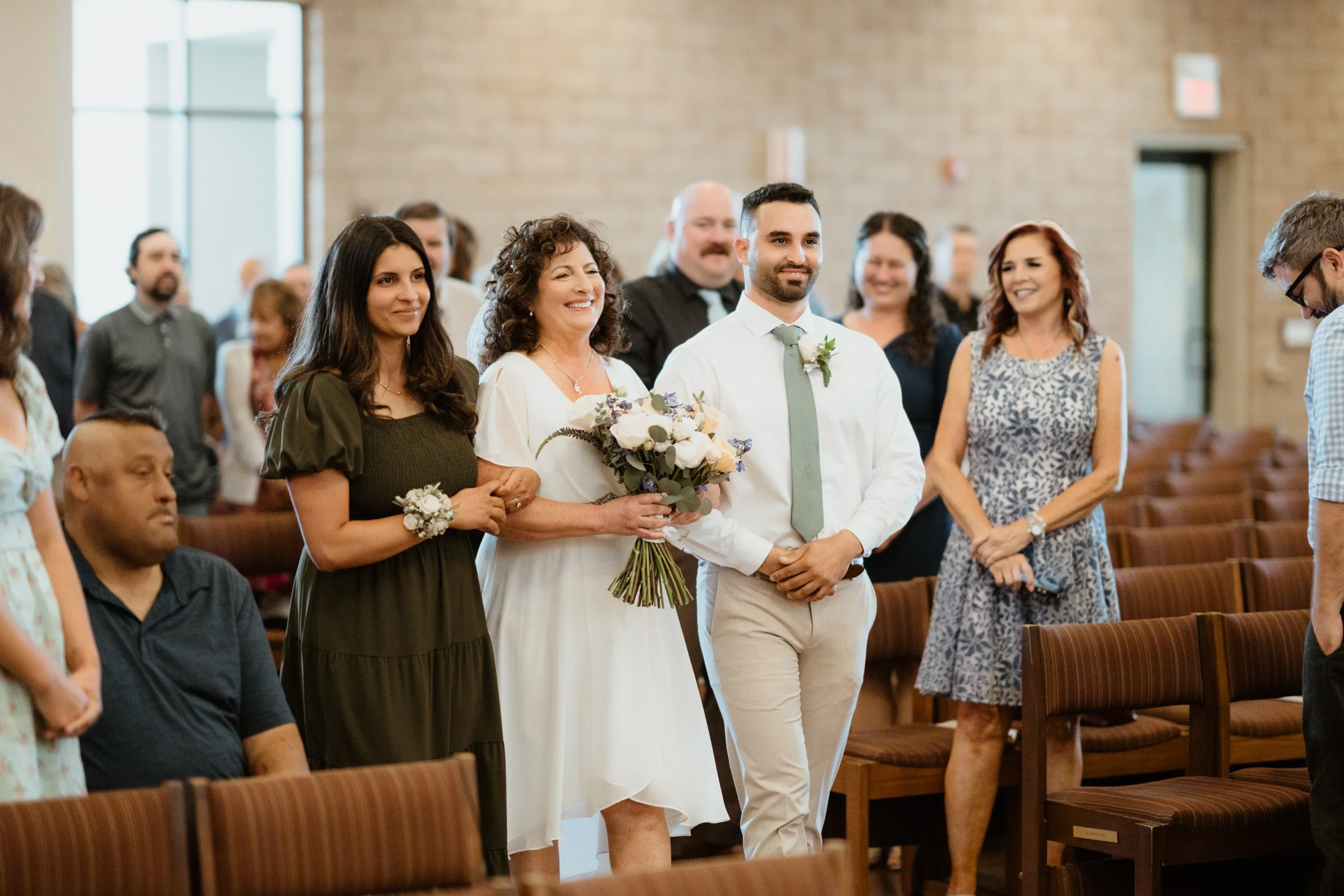 A bride in a white dress is being escorted down the aisle with her mother and a groom at a wedding ceremony in a church.
