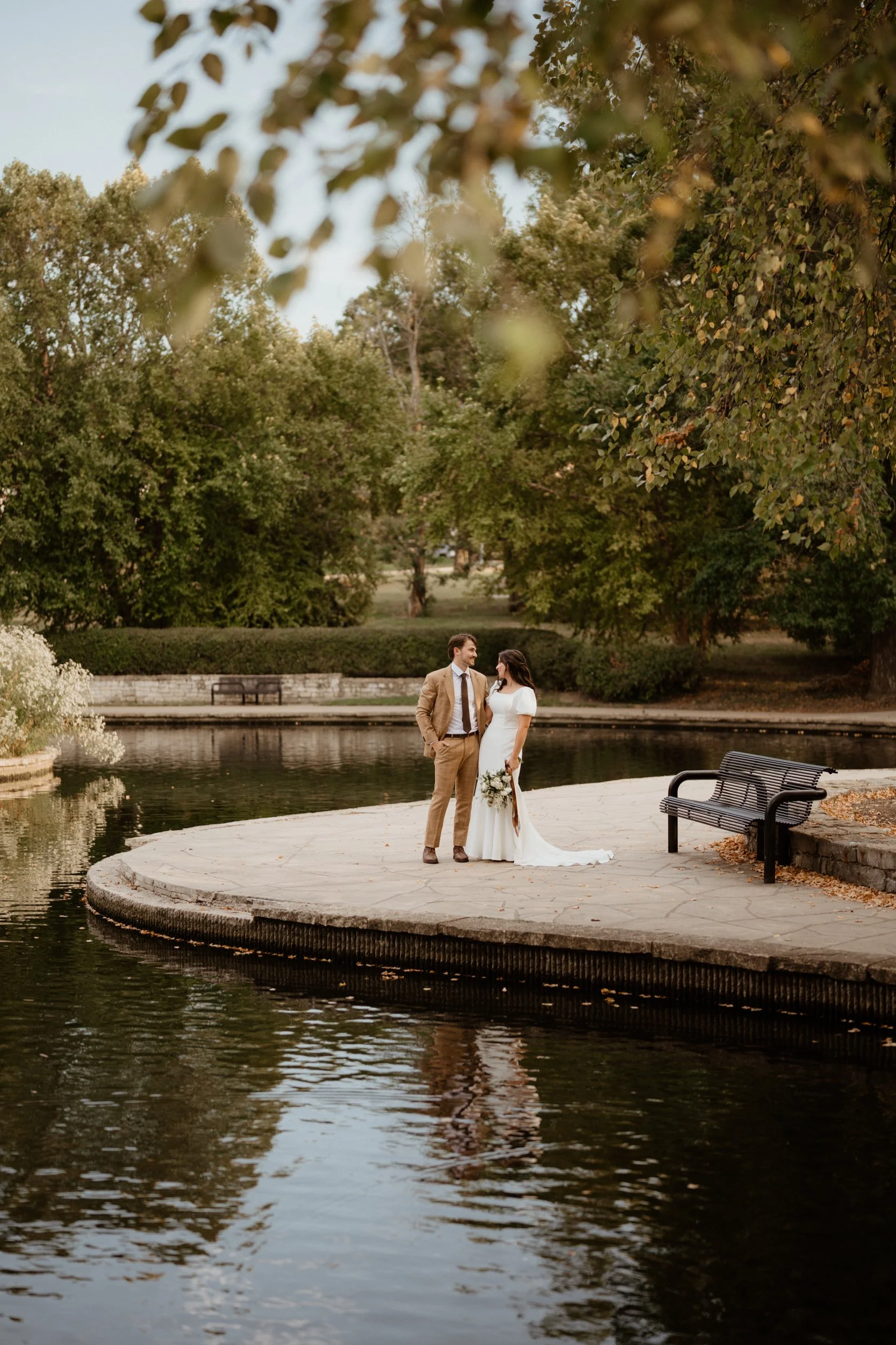 A bride and groom stand on a stone platform over a small pond in a park, surrounded by green trees, during a wedding photoshoot.