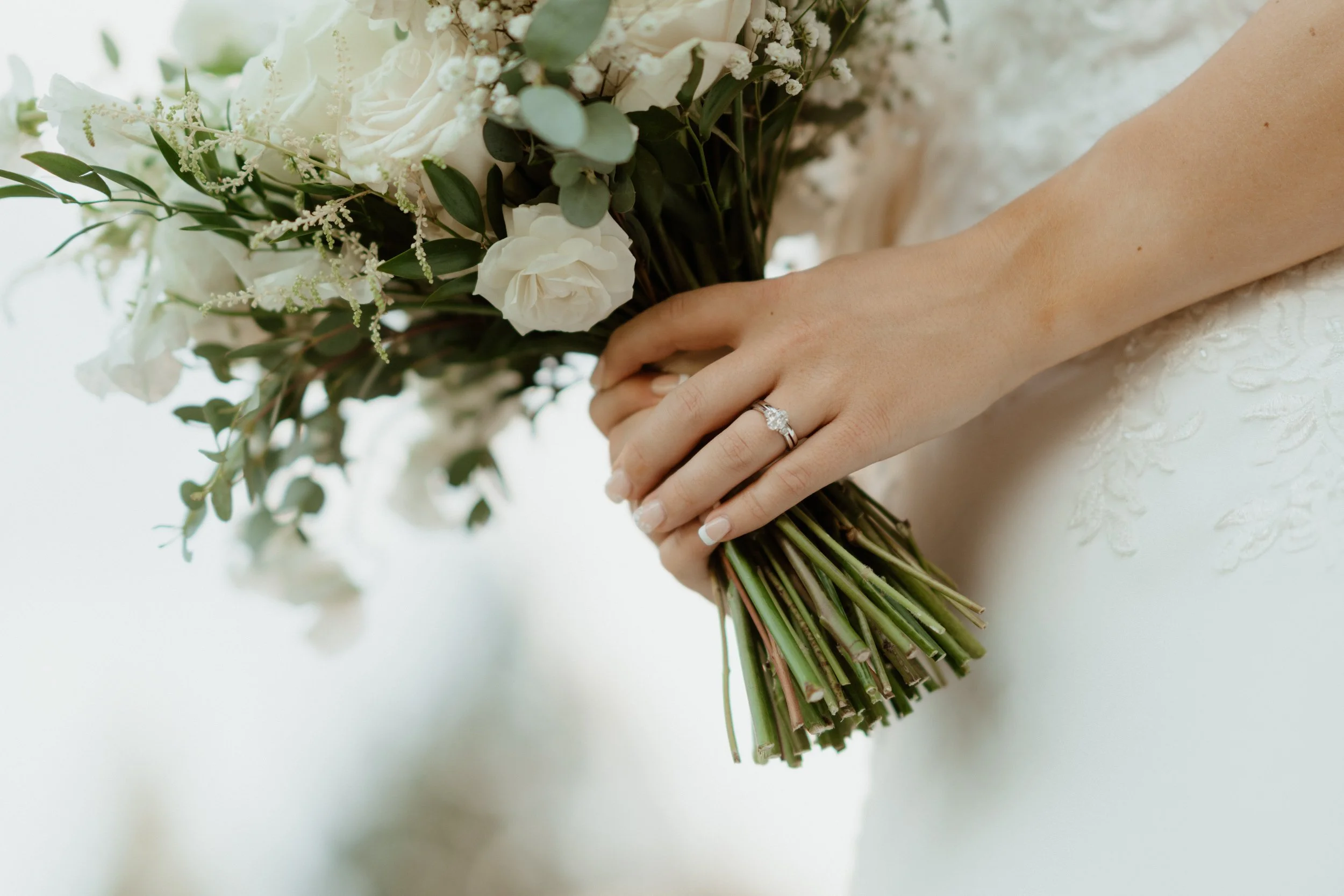 Close-up of a bride's hand holding a bouquet of white flowers, showing an engagement ring on her finger.