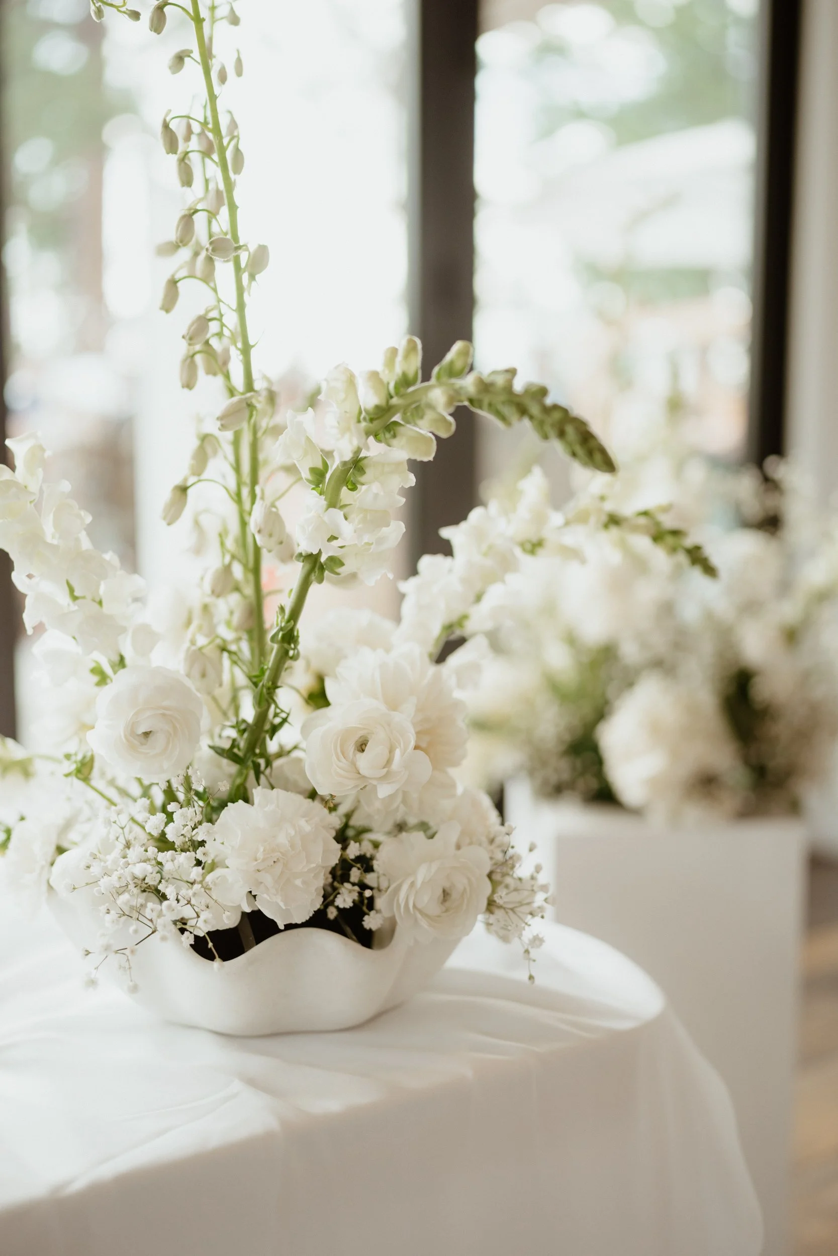 White floral arrangement in a decorative vase on a table with a white tablecloth, in front of a window.
