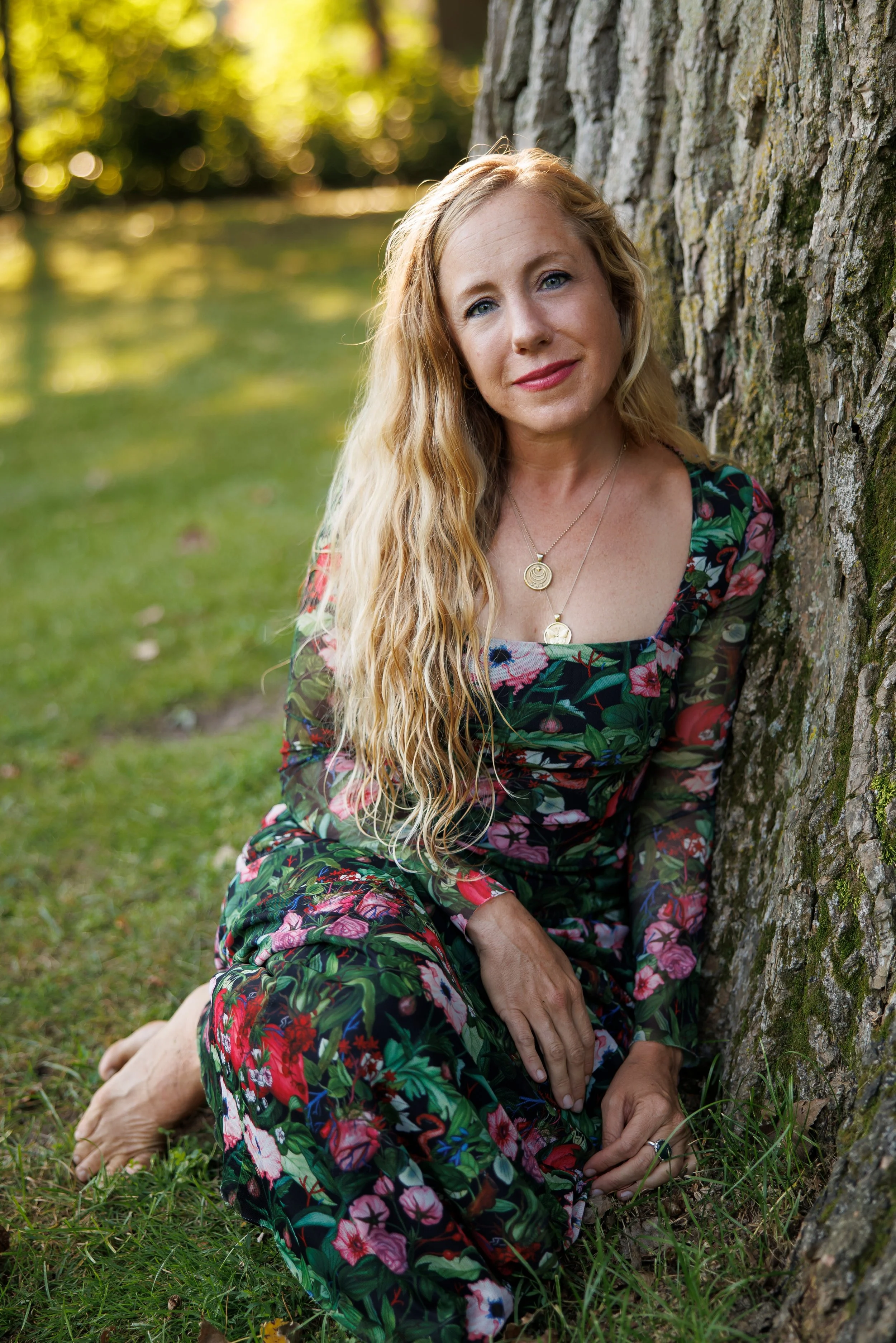 A woman with long, wavy blonde hair sitting outdoors against a tree trunk, wearing a floral dress and layered gold necklaces, looking at the camera.