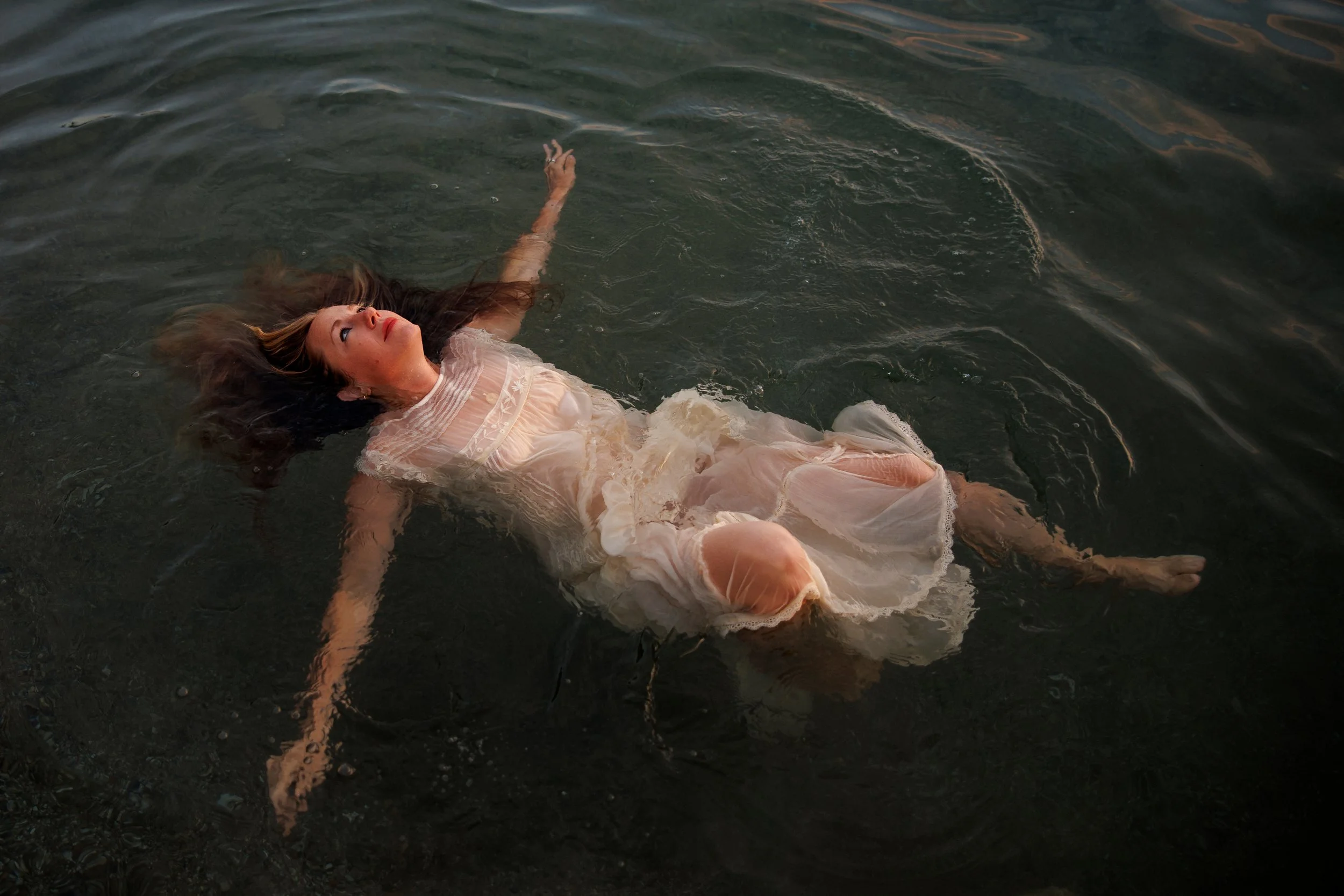 A woman lying on her back in dark water, wearing a light-colored dress, with her arms outstretched and her hair spread around her head.