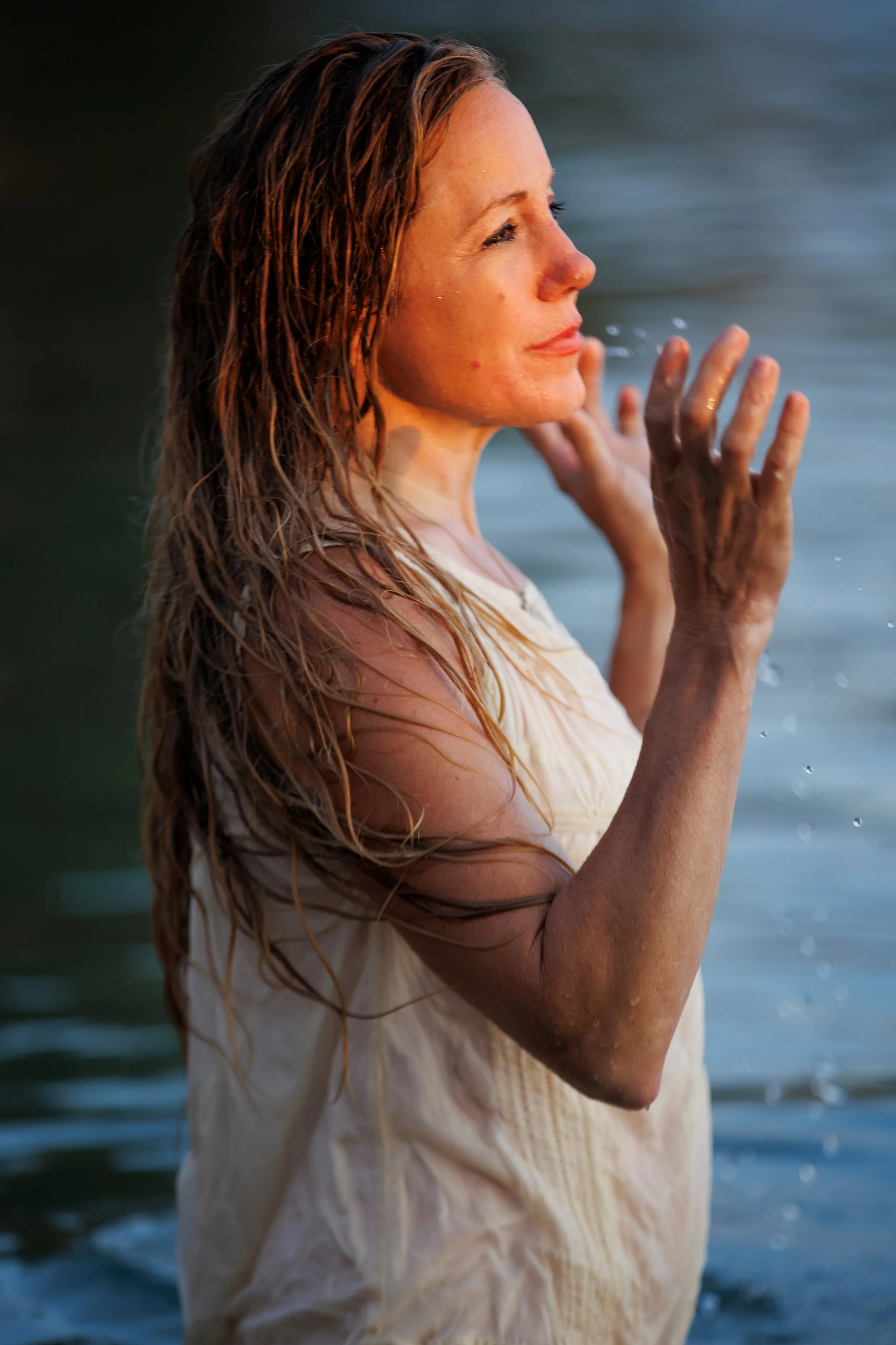 A woman with wet hair in a white dress standing in a body of water, facing sideways, with her hands near her face.