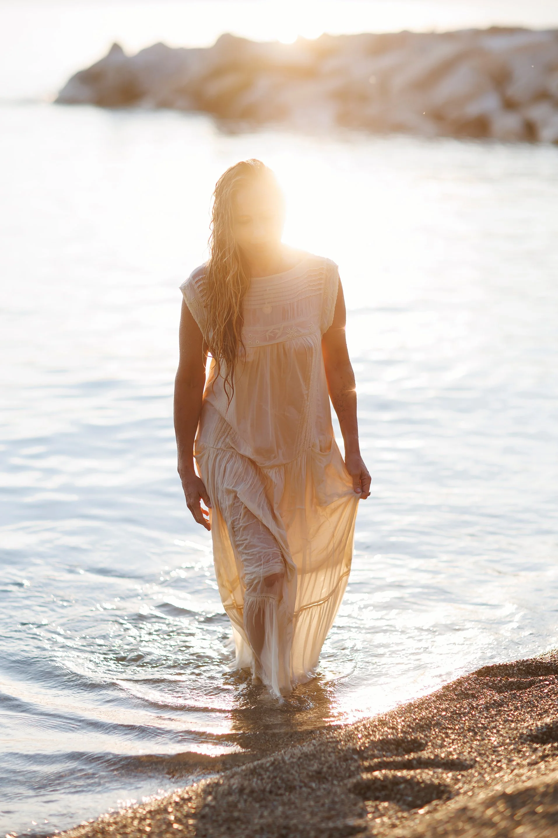 A woman in a white dress walking in shallow water at the beach during sunset.