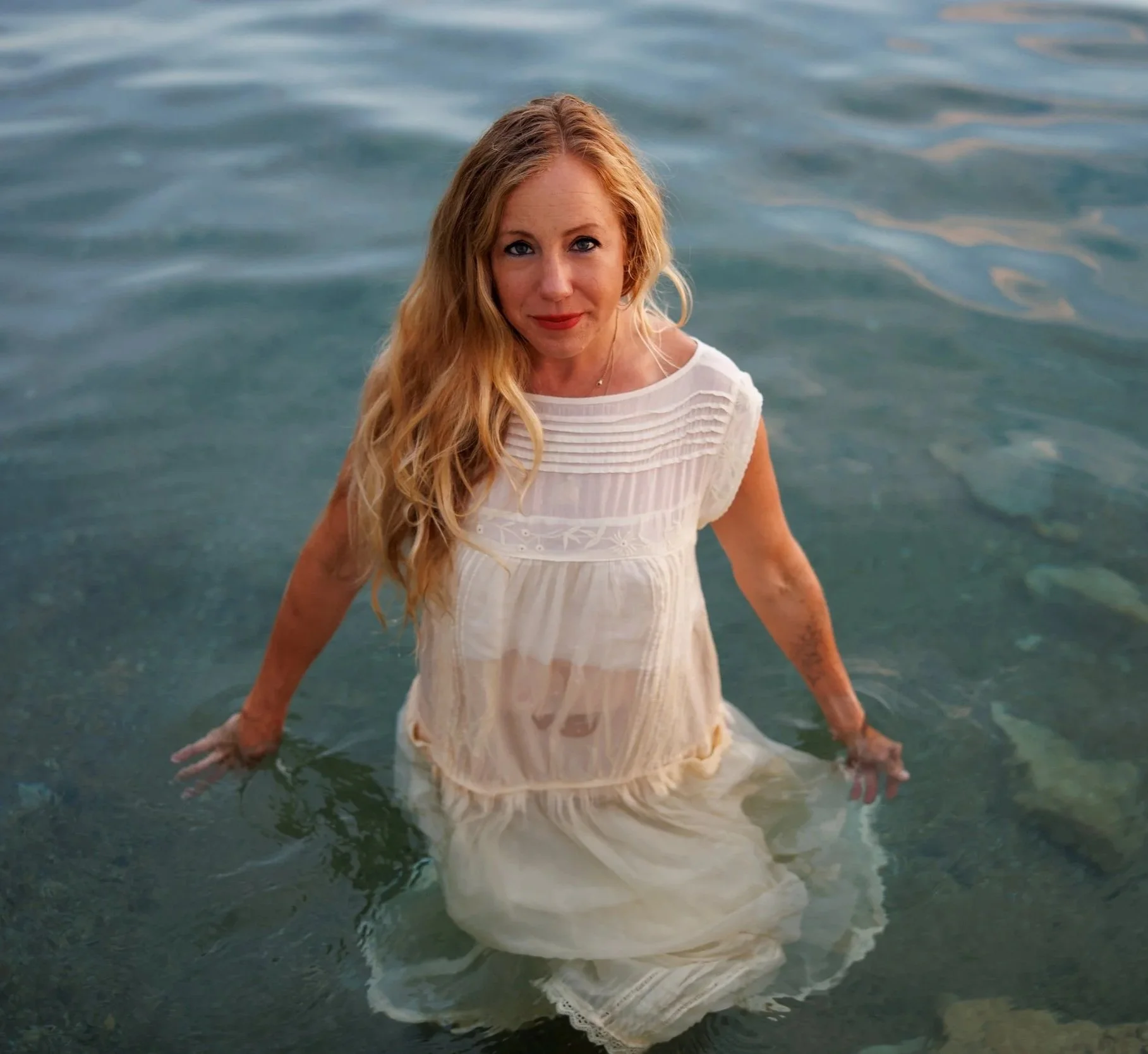 A woman with long blonde hair in a white dress standing in shallow water at the beach.