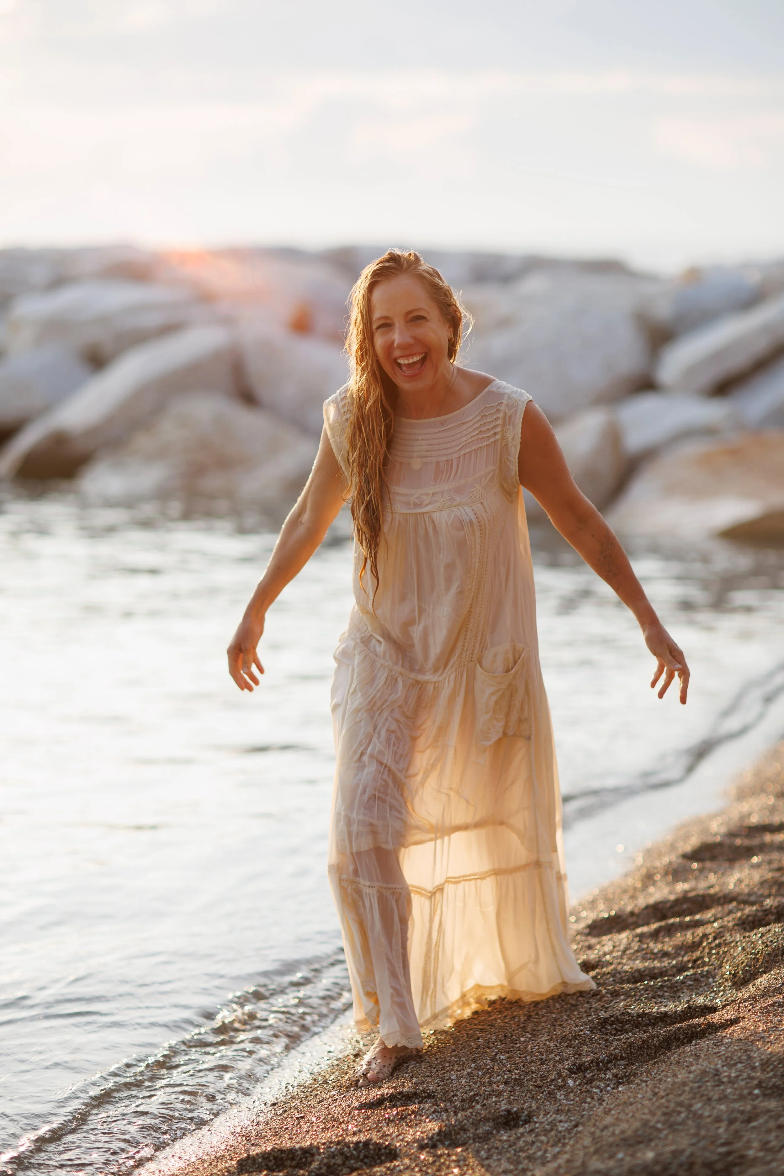 A woman in a light-colored, flowy dress with wet hair, smiling and walking at the edge of the water on a sandy beach during sunset.