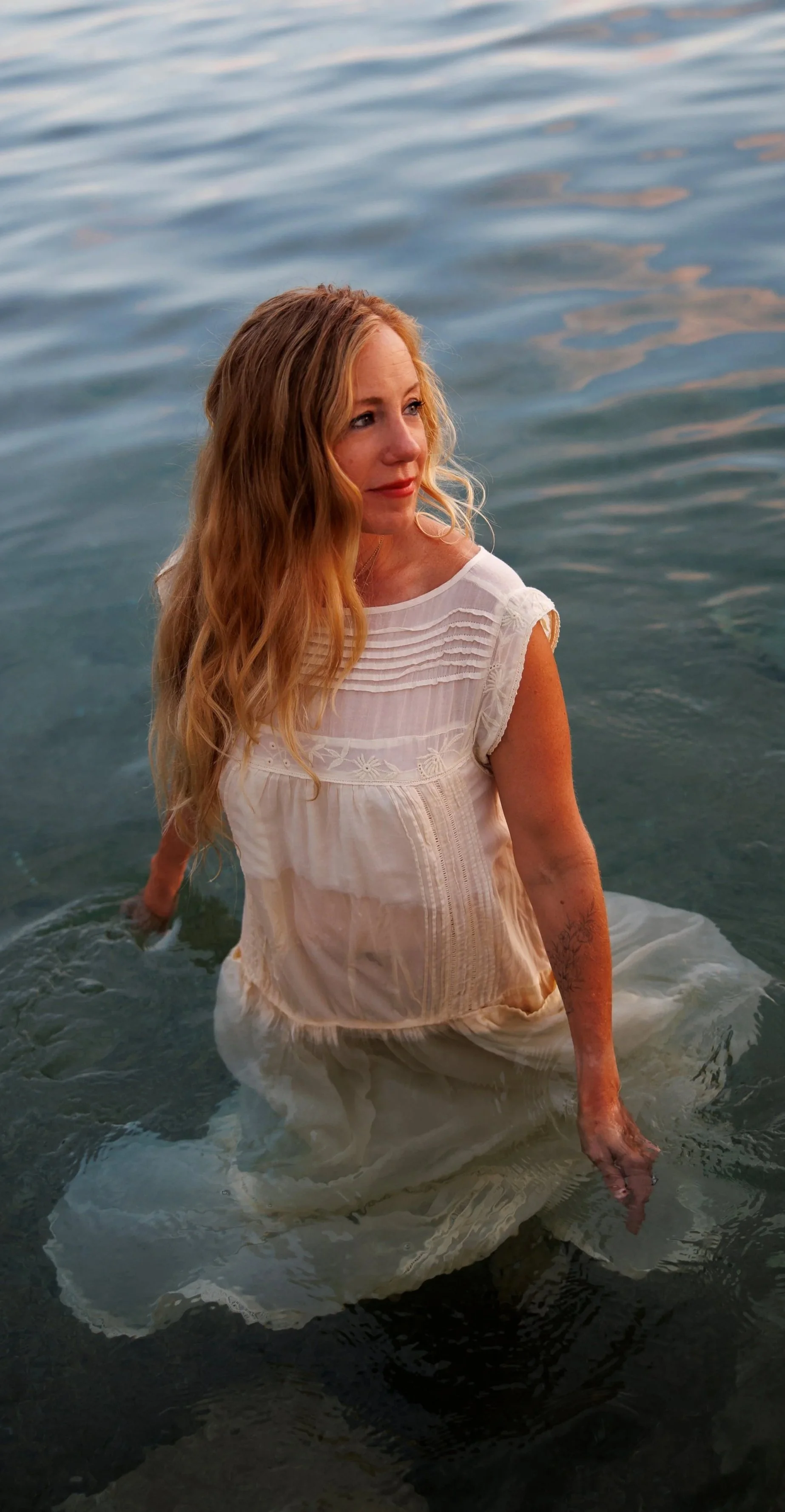 A woman with long wavy red hair wearing a white dress, standing in water with a calm expression, during sunset.