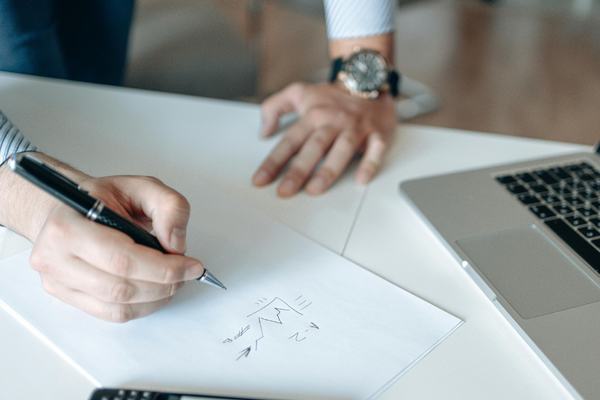 Someone is writing on a piece of paper with a pen, with a laptop and another person's hand resting nearby on a white table.