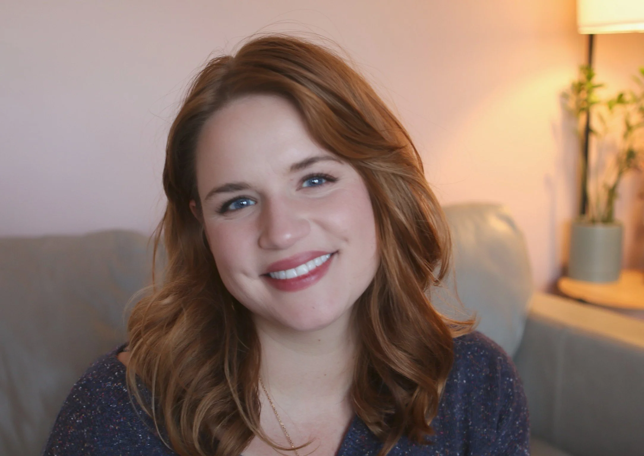 A young woman with red hair and blue eyes smiling while sitting on a beige couch in a cozy living room, with a lamp and potted plant in the background.