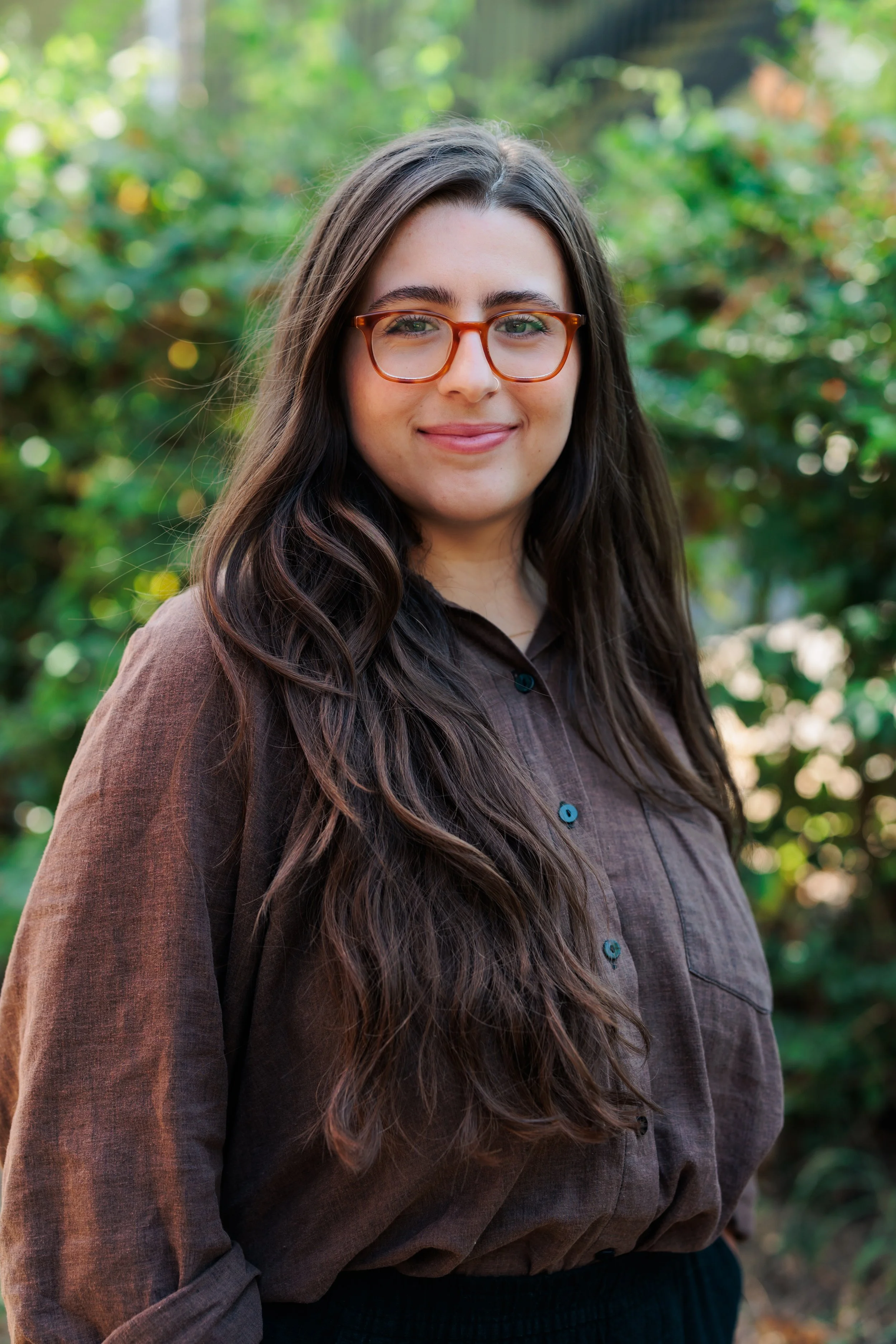 A woman with long dark hair, wearing glasses and a brown shirt, standing outdoors with greenery in the background.