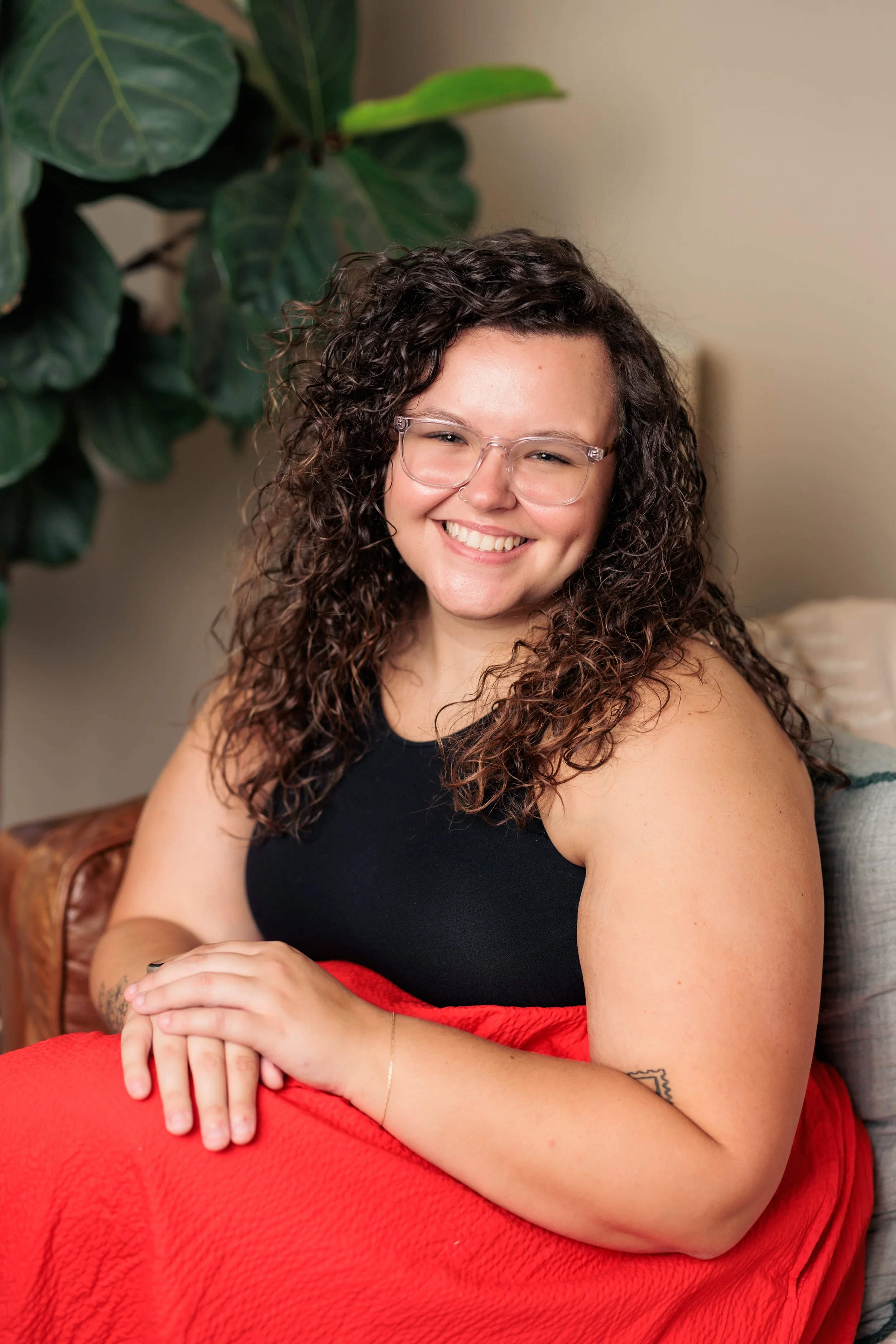 A woman with curly brown hair, glasses, and a black sleeveless top smiling, sitting on a couch with a green plant in the background.