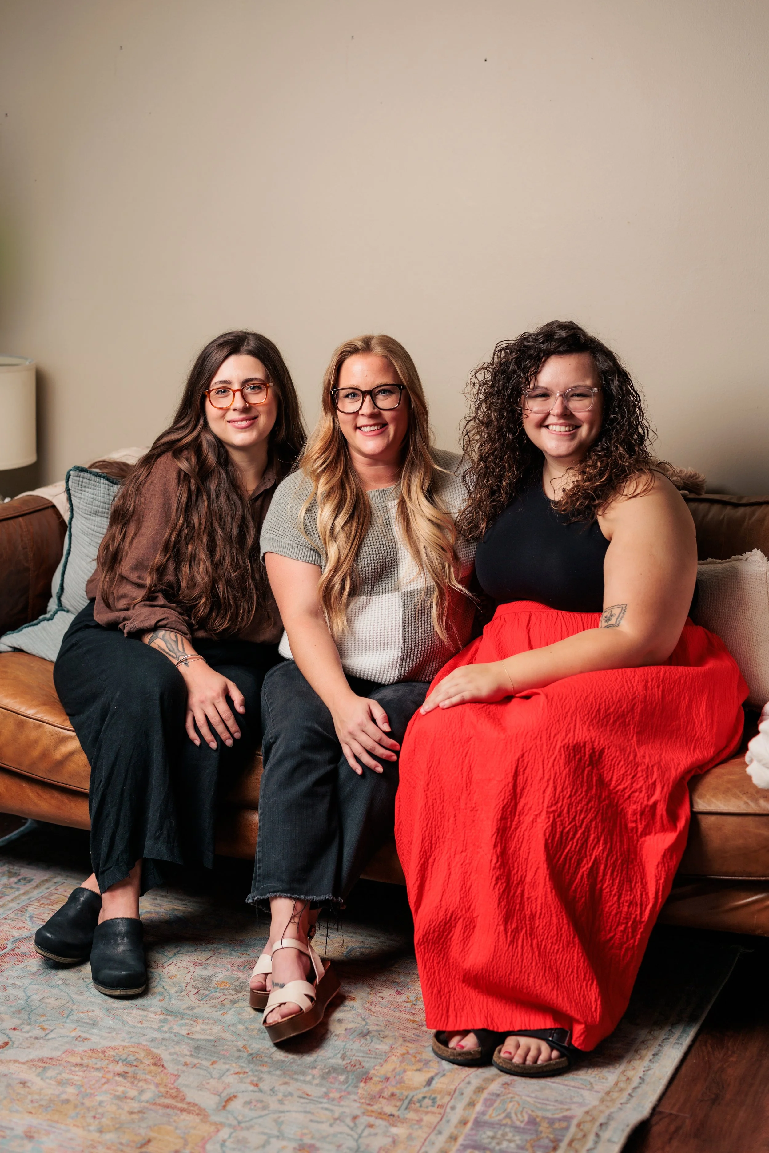 Three women, therapists sitting on a brown leather couch in a living room, smiling at the camera.