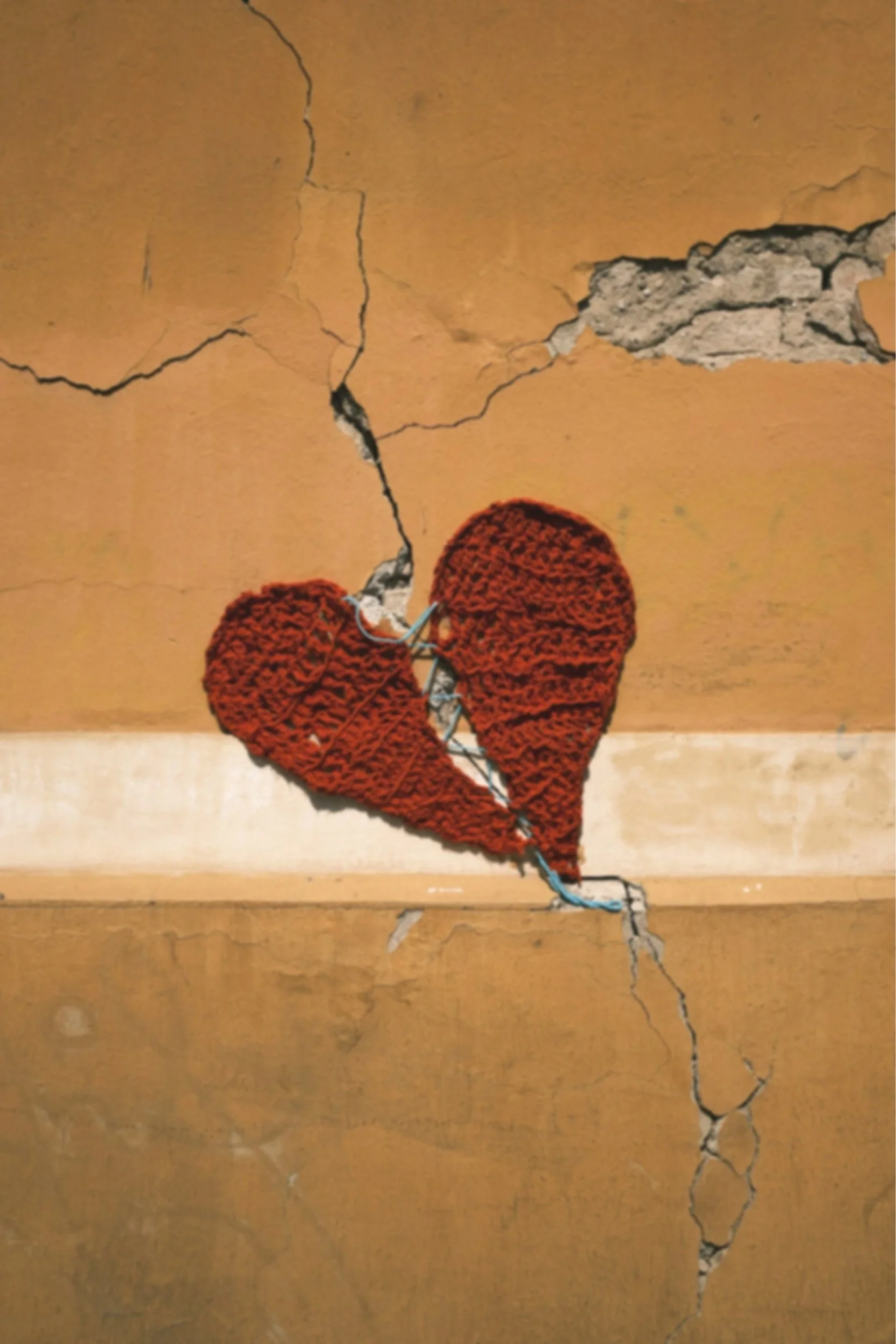 A broken, knitted red heart mural on a cracked yellow wall.