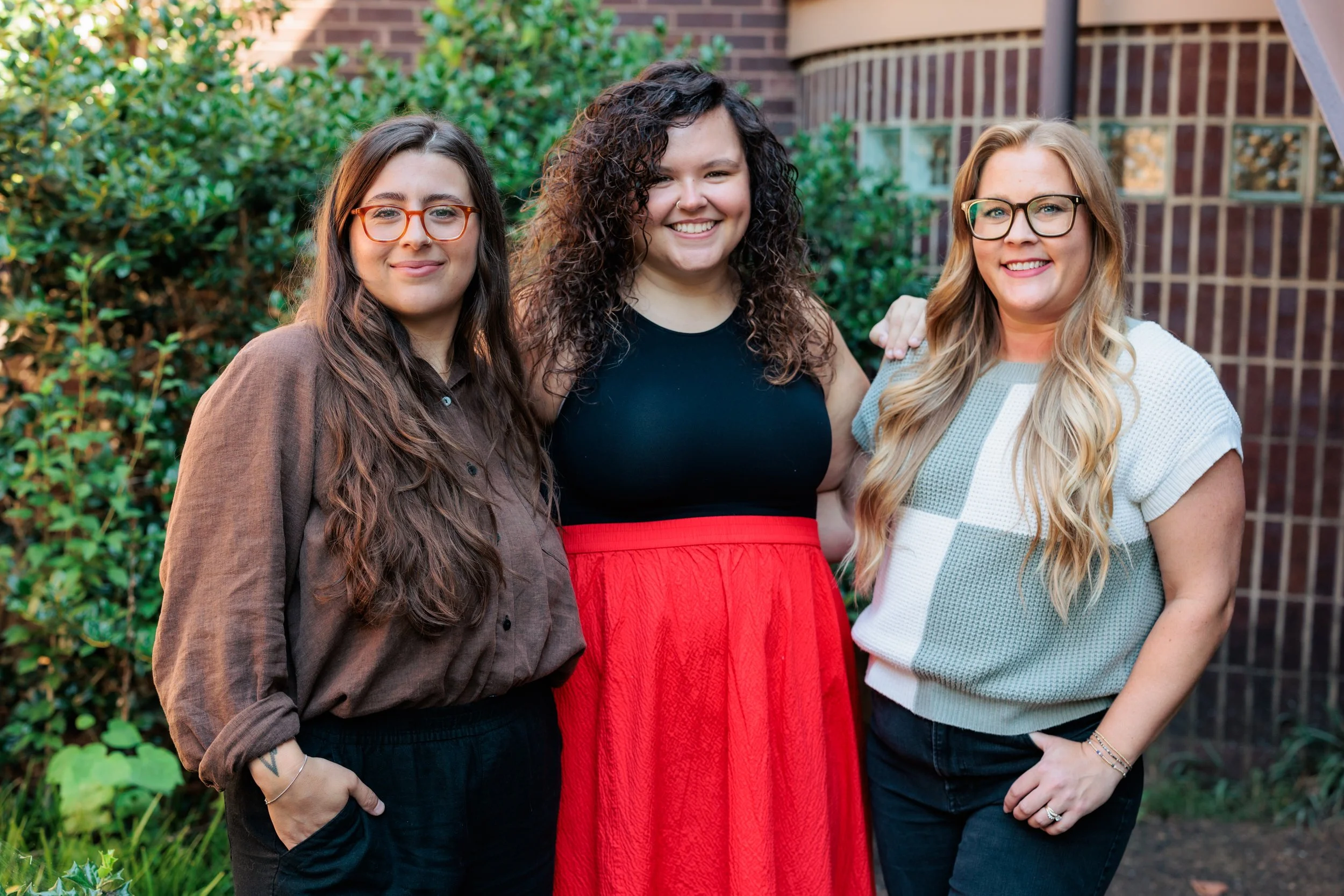 Three women standing outdoors in front of green bushes and a brick building, smiling at the camera.