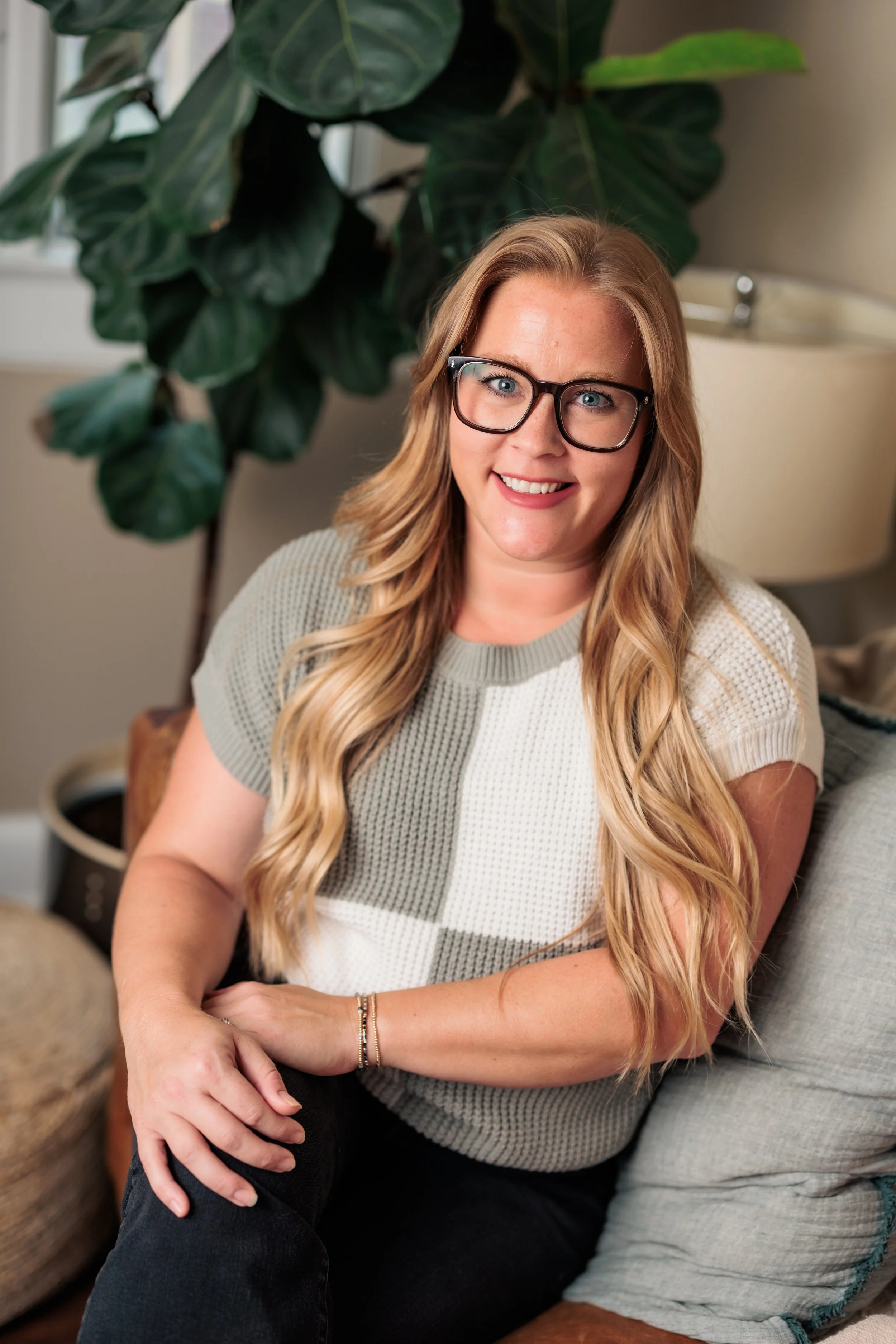 A woman with long blonde hair, glasses, and a checkered sweater sitting on a sofa, smiling at the camera with a houseplant and a lamp in the background.