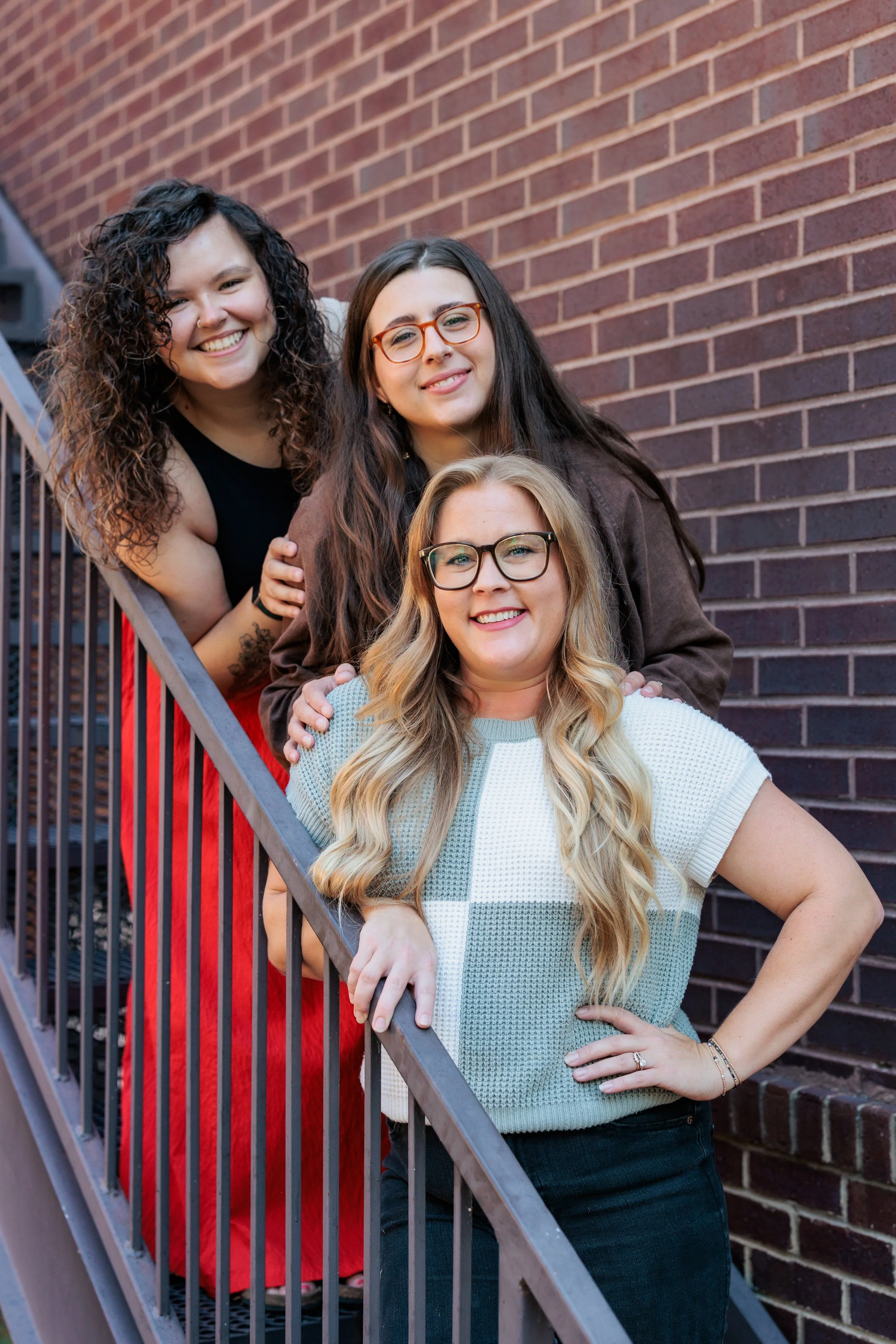 Four women smiling on a staircase against a brick wall, all wearing casual clothing.