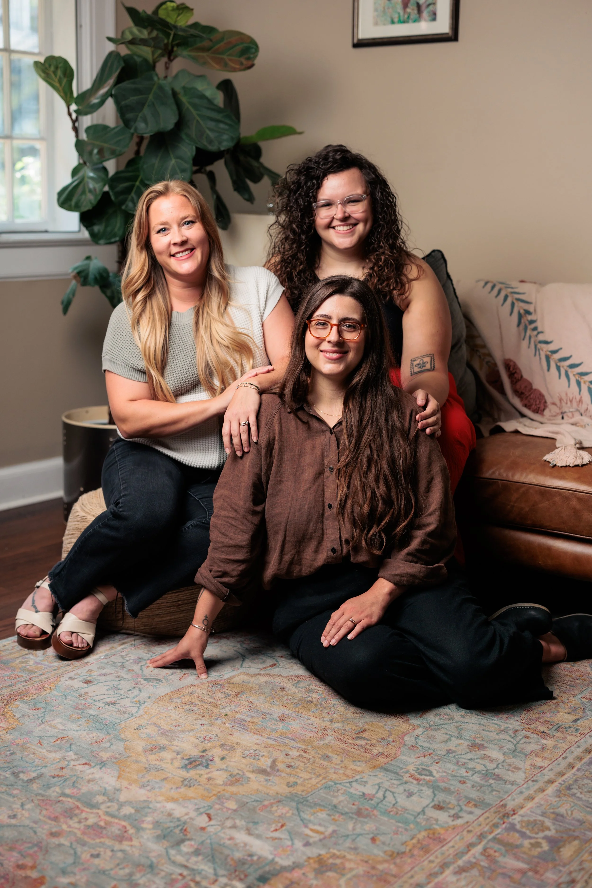 Four women smiling and posing together in a cozy room with a large plant, framed picture, a window, and a patterned rug.
