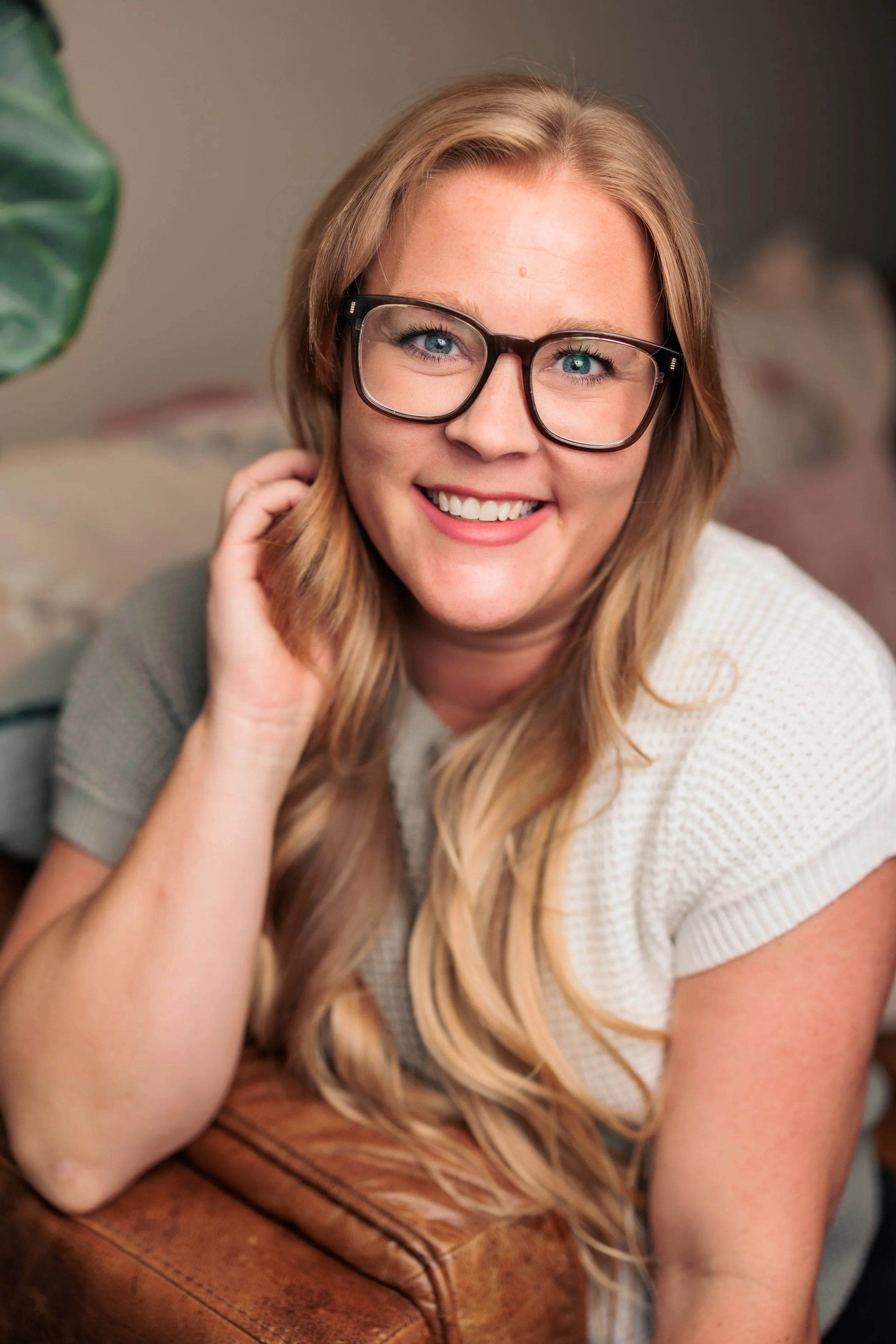 A woman with long blonde hair, wearing glasses and a white shirt, smiling at the camera indoors.