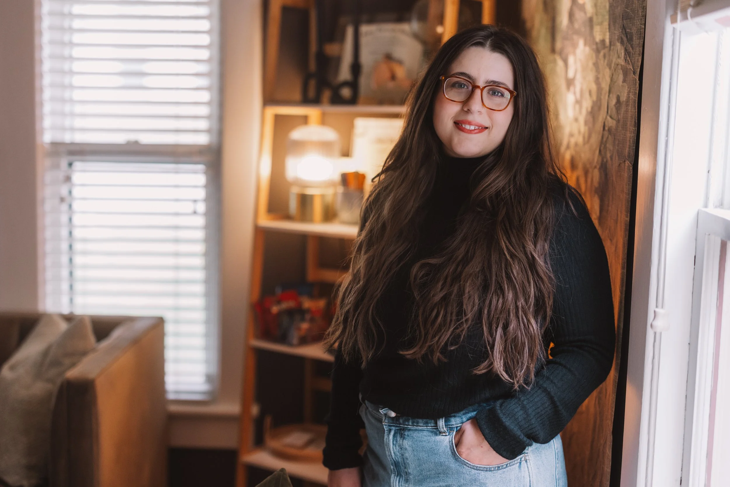 A young woman with long wavy brown hair, wearing glasses, a black turtleneck sweater, and light blue jeans, leaning against a wooden wall by a window, smiling at the camera inside a cozy room with a shelving unit and warm lighting.