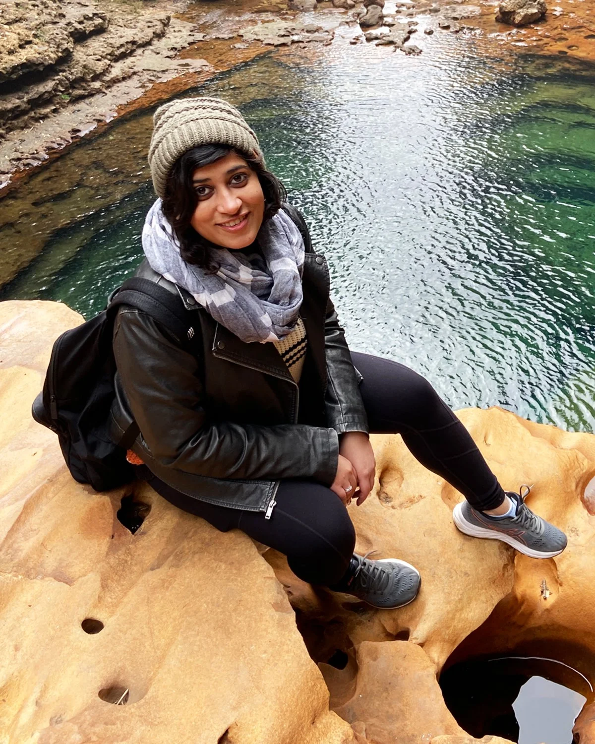 A woman sitting on a rock near a pond, wearing a gray beanie, black leather jacket, striped shirt, black pants, gray sneakers, and a gray scarf, with a black backpack on her back.