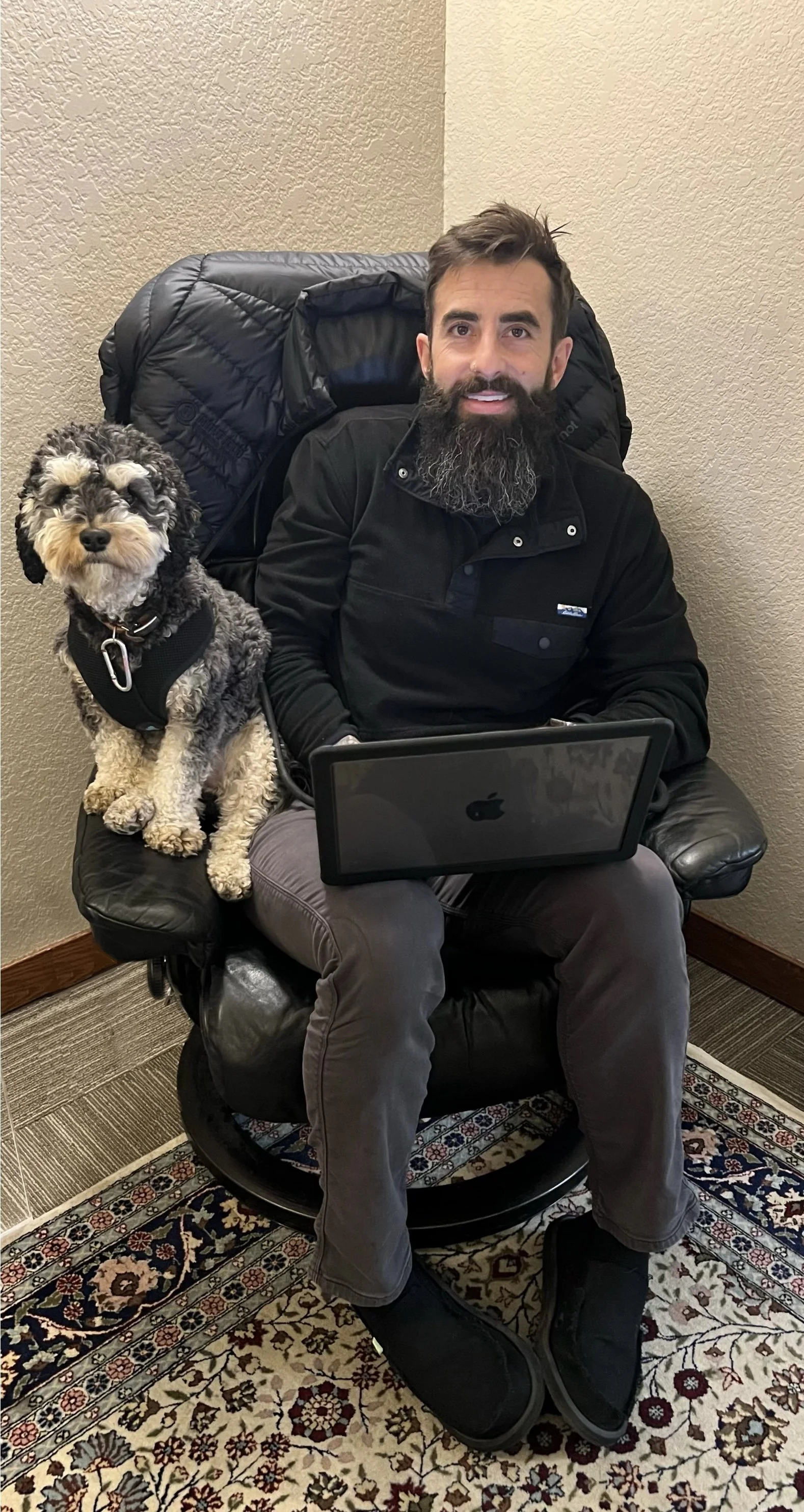 A man with a beard sitting in a black leather chair with a small black, white, and gray dog on his left side. The man has a laptop on his lap and a jacket on the back of the chair. The setting appears to be indoors with beige walls, a patterned rug, and a corner of a room visible.