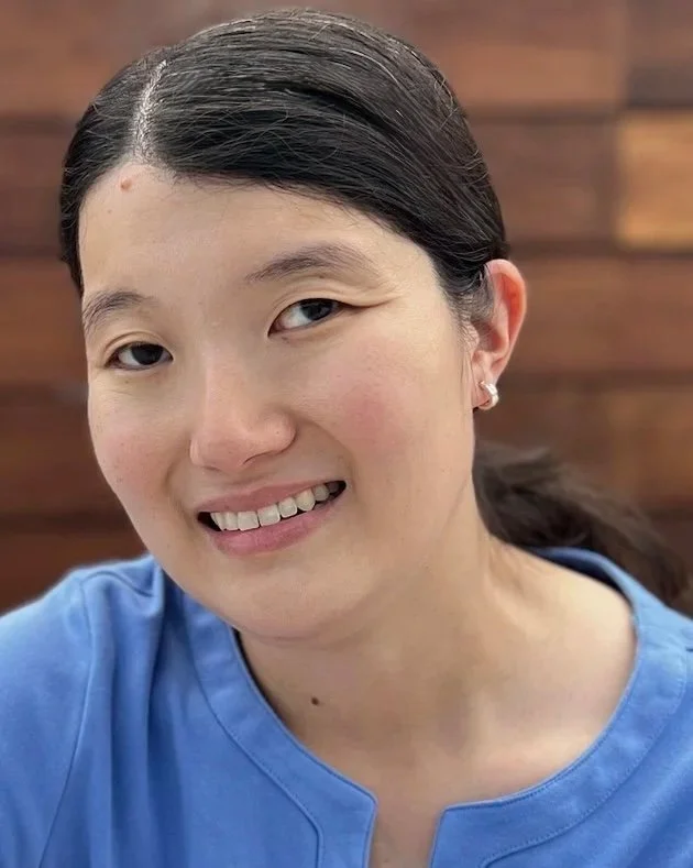 A woman with long dark hair pulled back, smiling slightly, wearing a blue top with a rounded neckline and small earrings, with a wooden wall in the background.