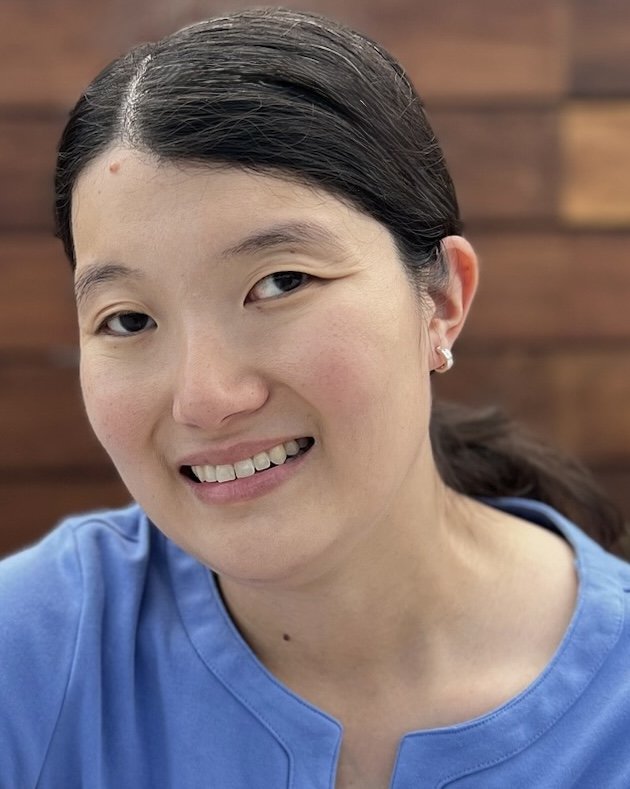 Close-up photo of a smiling woman with dark hair tied back, wearing a blue top and small earrings, against a blurred wooden background.