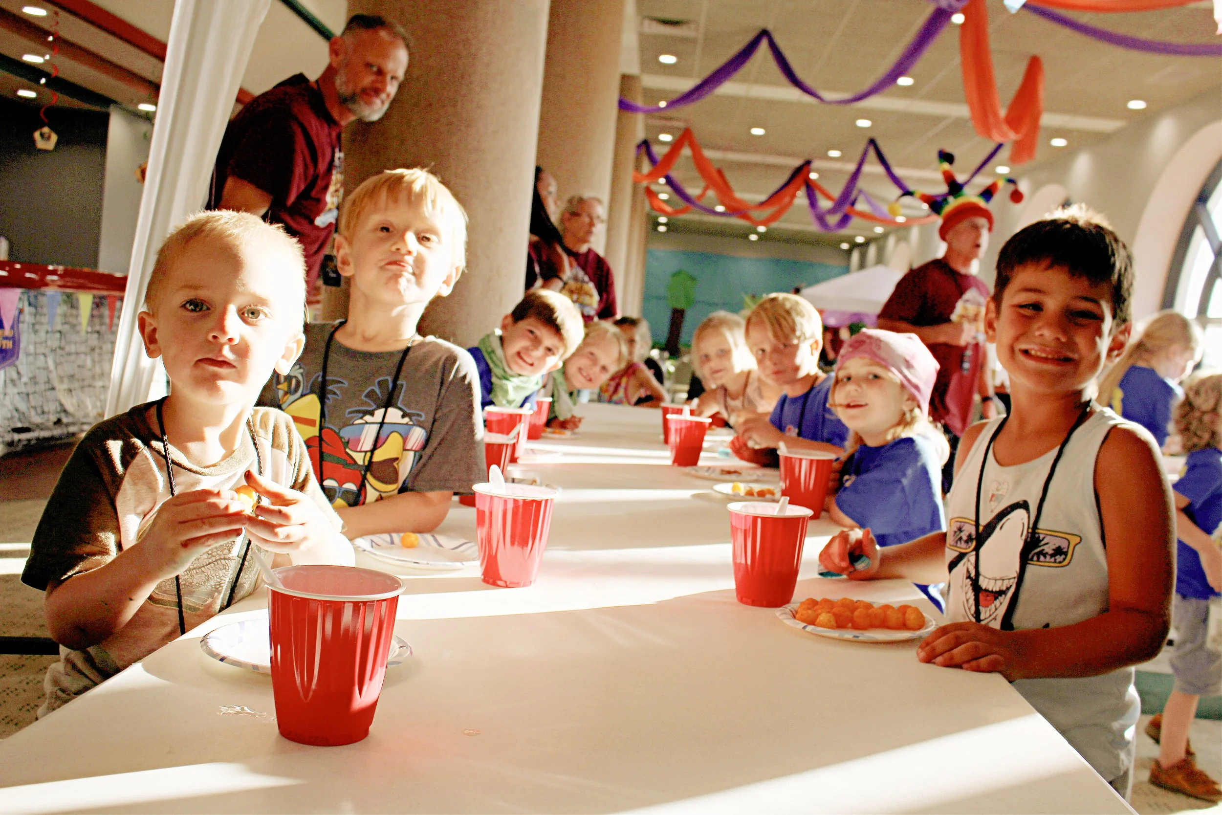Children and adults gather at a long table decorated with orange and purple streamers, celebrating a party. Kids are seated with red cups and plates of snacks in a festively decorated room with purple and orange streamers overhead.