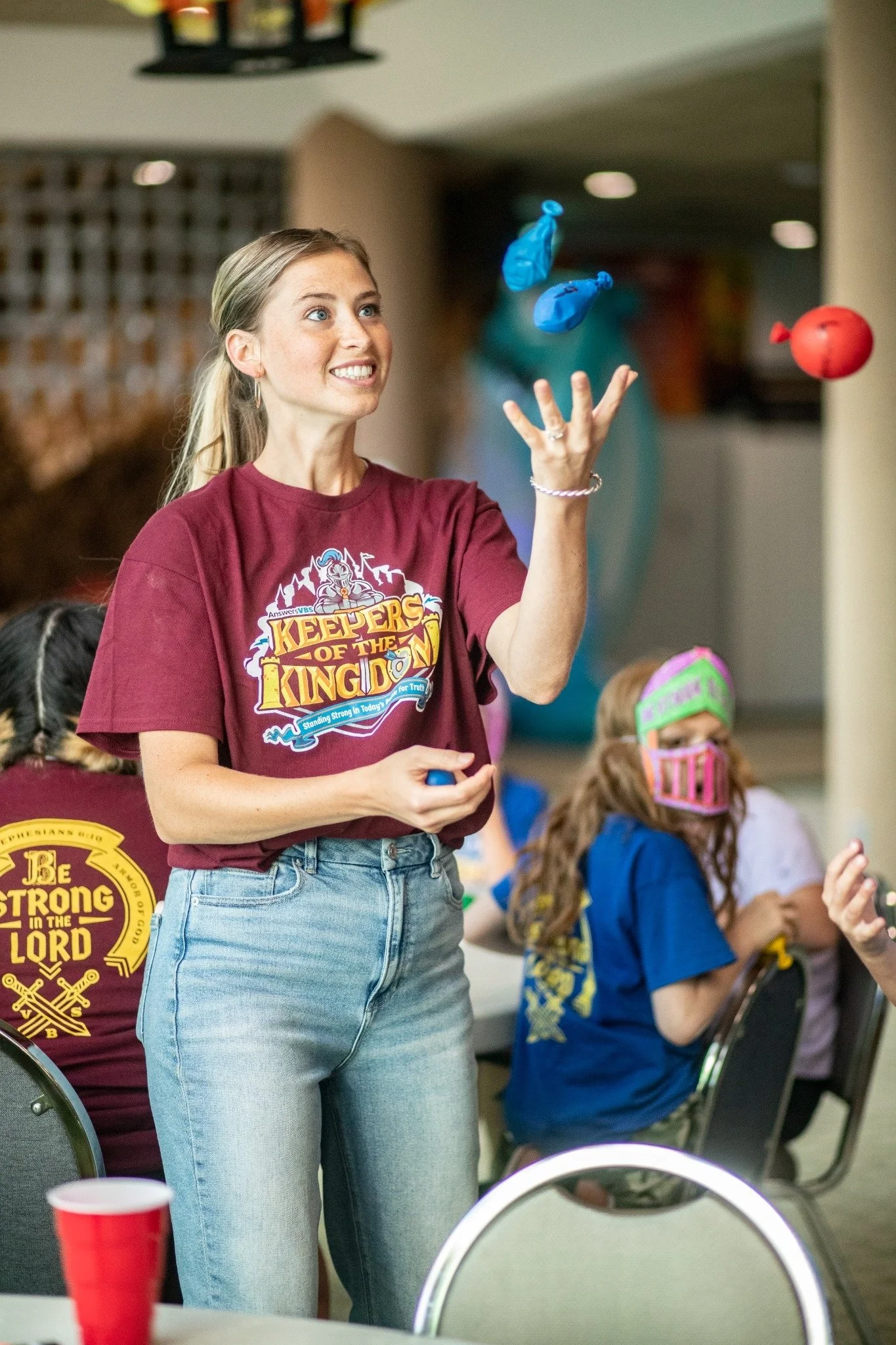 A young woman with blonde hair in a ponytail, wearing a maroon t-shirt with a cartoon character and text that says 'Keepers of the Kingdom,' is juggling blue and red balls in a room with children and chairs. The background includes children sitting at tables, some wearing colorful face masks, and a yellow sign that says 'Be Strong in the Lord.'