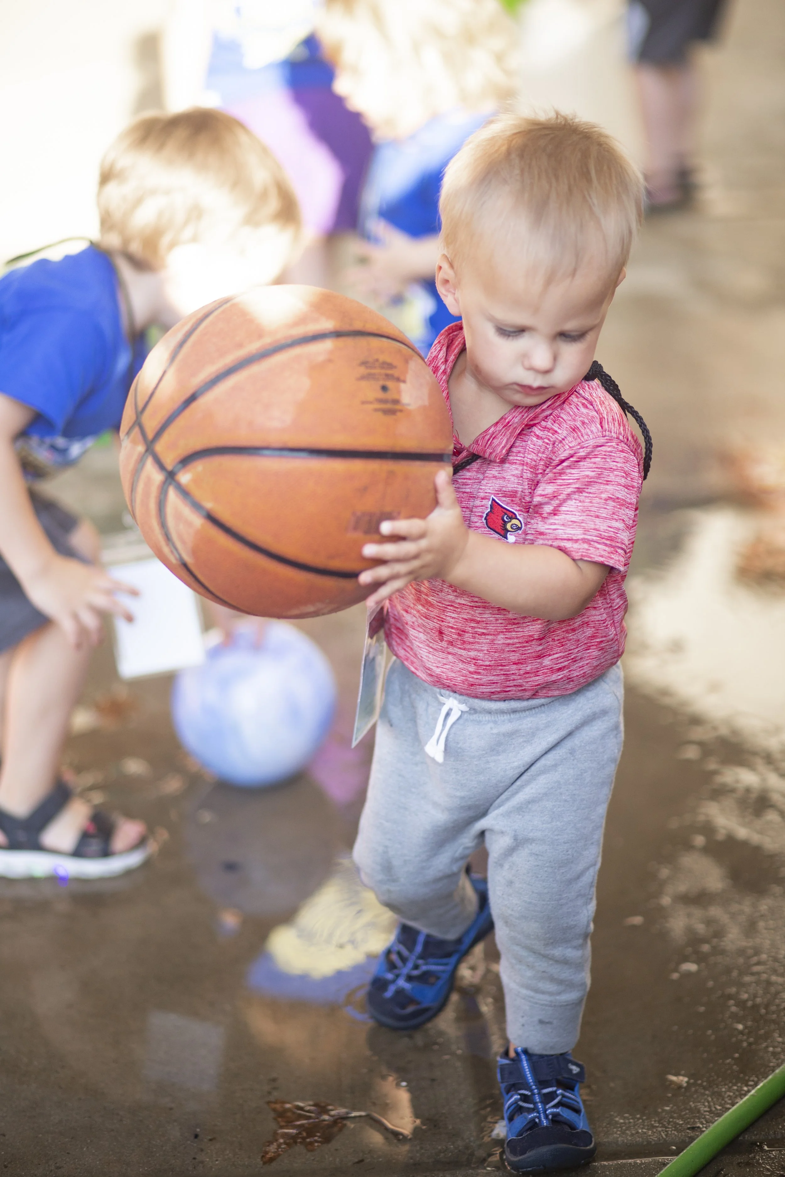 Kids playing with a basketball indoors. Focus on a young boy in a red shirt holding a basketball. Other children are visible in the background.