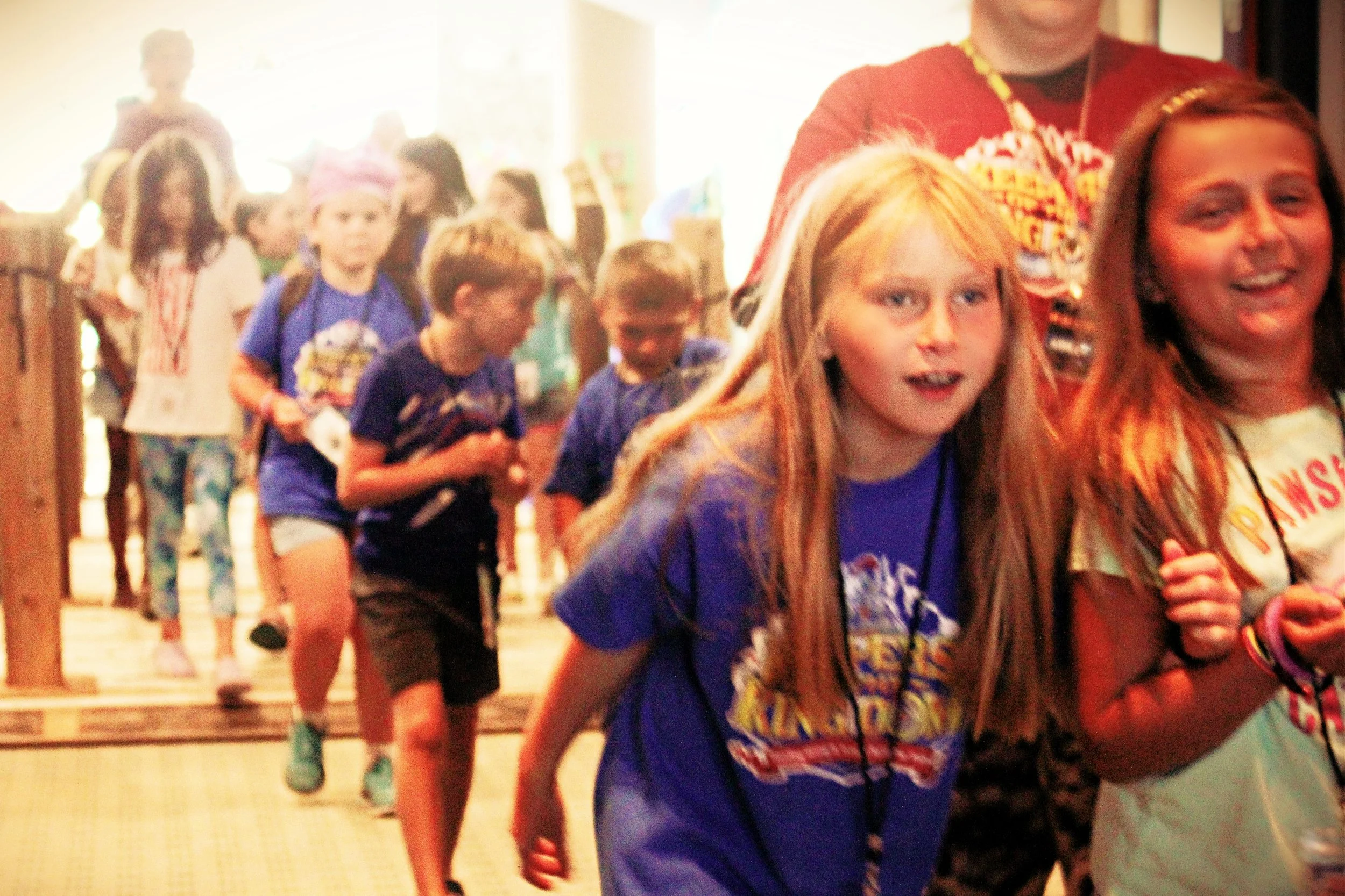 Children standing in line indoors, some wearing blue shirts and backpacks, with one girl in the front smiling, in a busy, brightly lit setting.